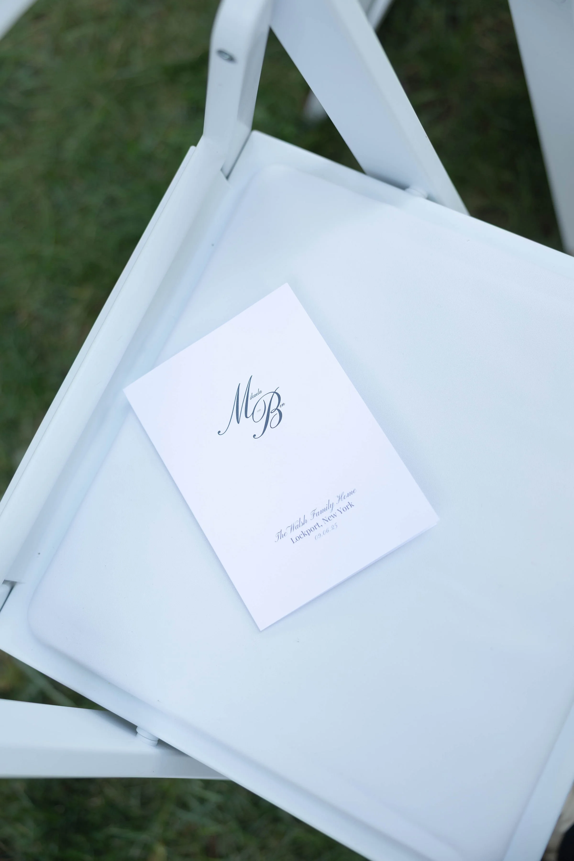Close-up of a white folding chair with a wedding program resting on the seat. The program has elegant black lettering and details about a wedding event.