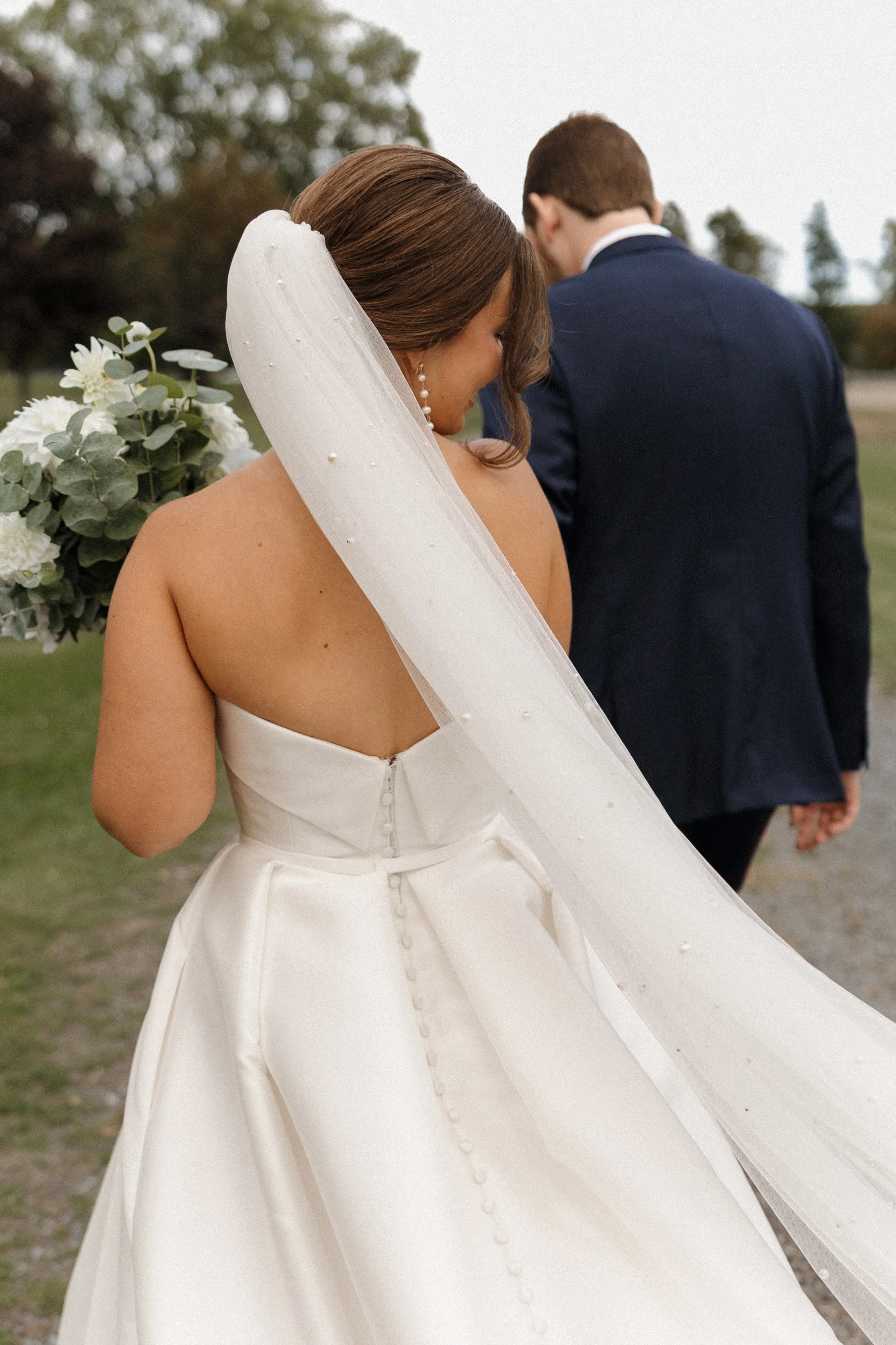 A bride in a strapless white wedding dress with a bow at the waist, wearing a veil with pearl accents, standing outdoors with her head bowed and eyes closed. A bouquet of white flowers and green foliage is visible in the background. Behind her, a gro