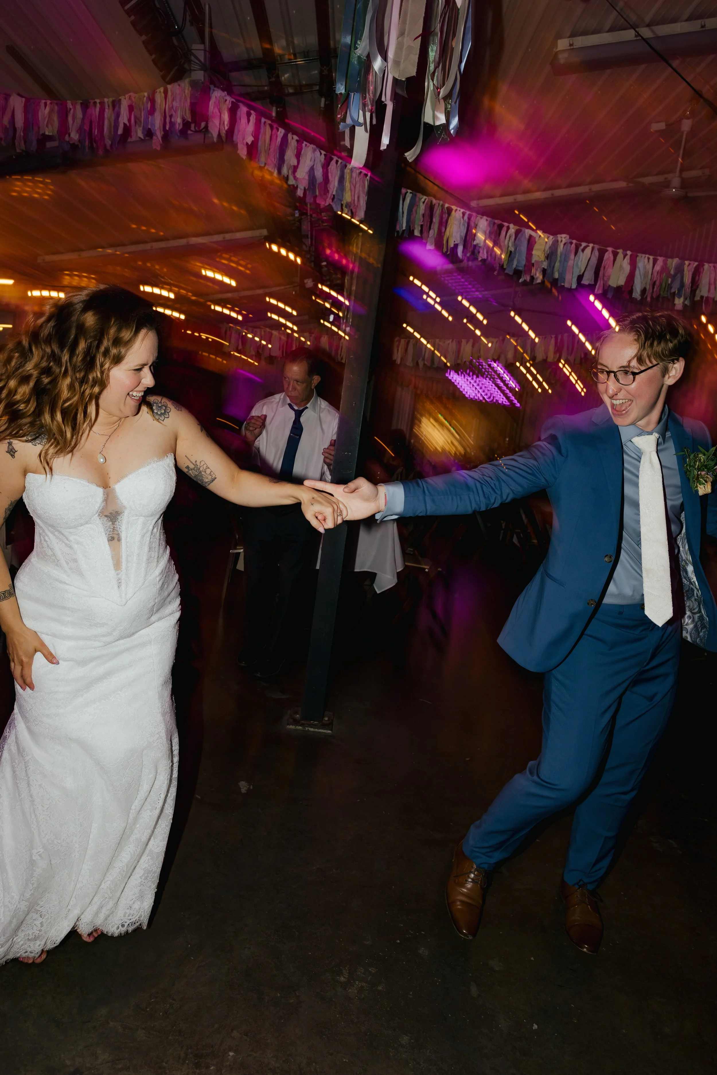 A woman in a white lace wedding dress holding hands with a man in a blue suit and glasses, dancing at a celebration with colorful hanging decorations and purple lighting.