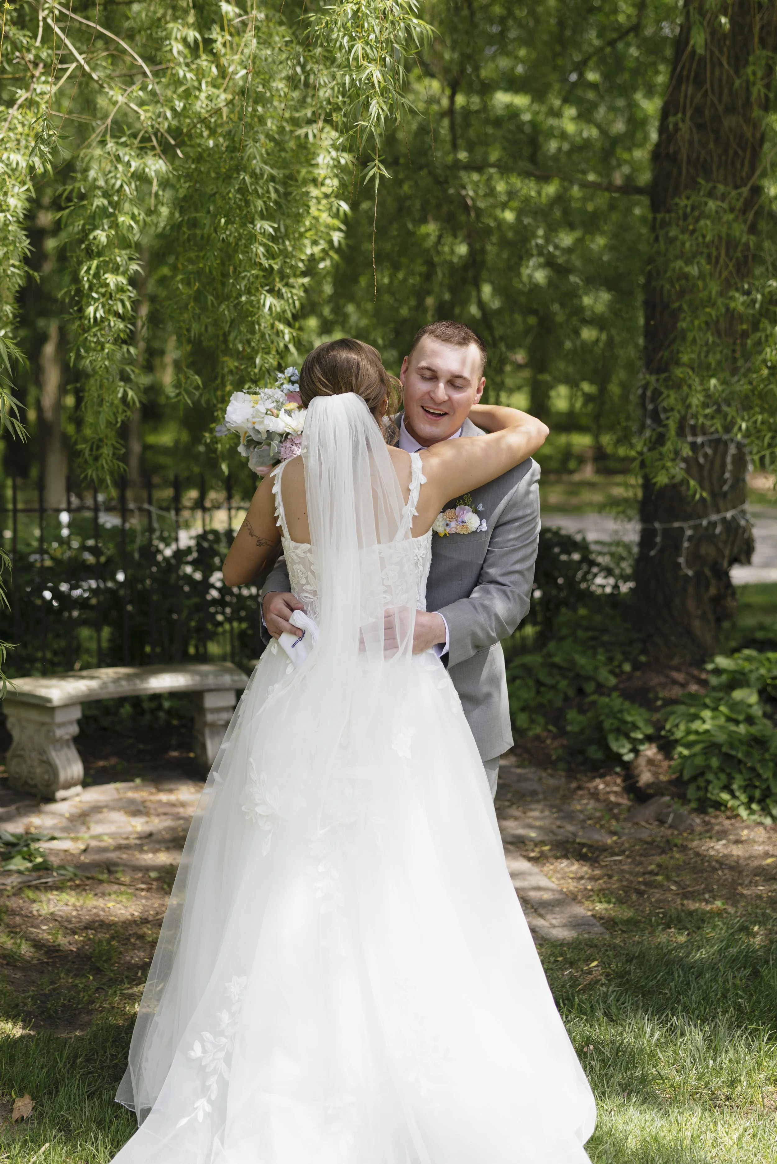A wedding couple embracing outdoors surrounded by green trees and plants, with a bench nearby and dappled sunlight.