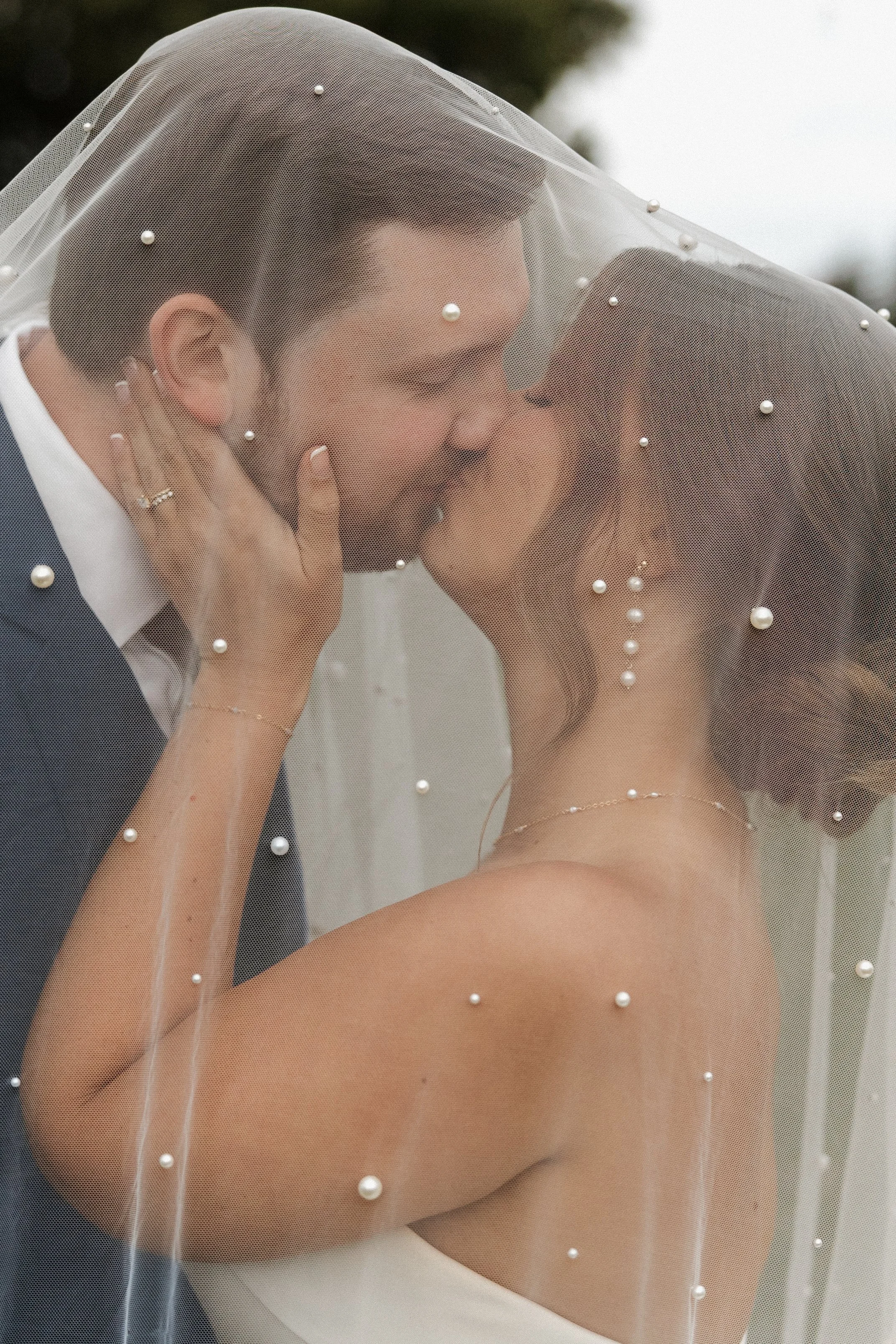 A bride and groom sharing a kiss under a sheer veil decorated with pearls, with the bride's face partially visible and the groom's dark hair showing.