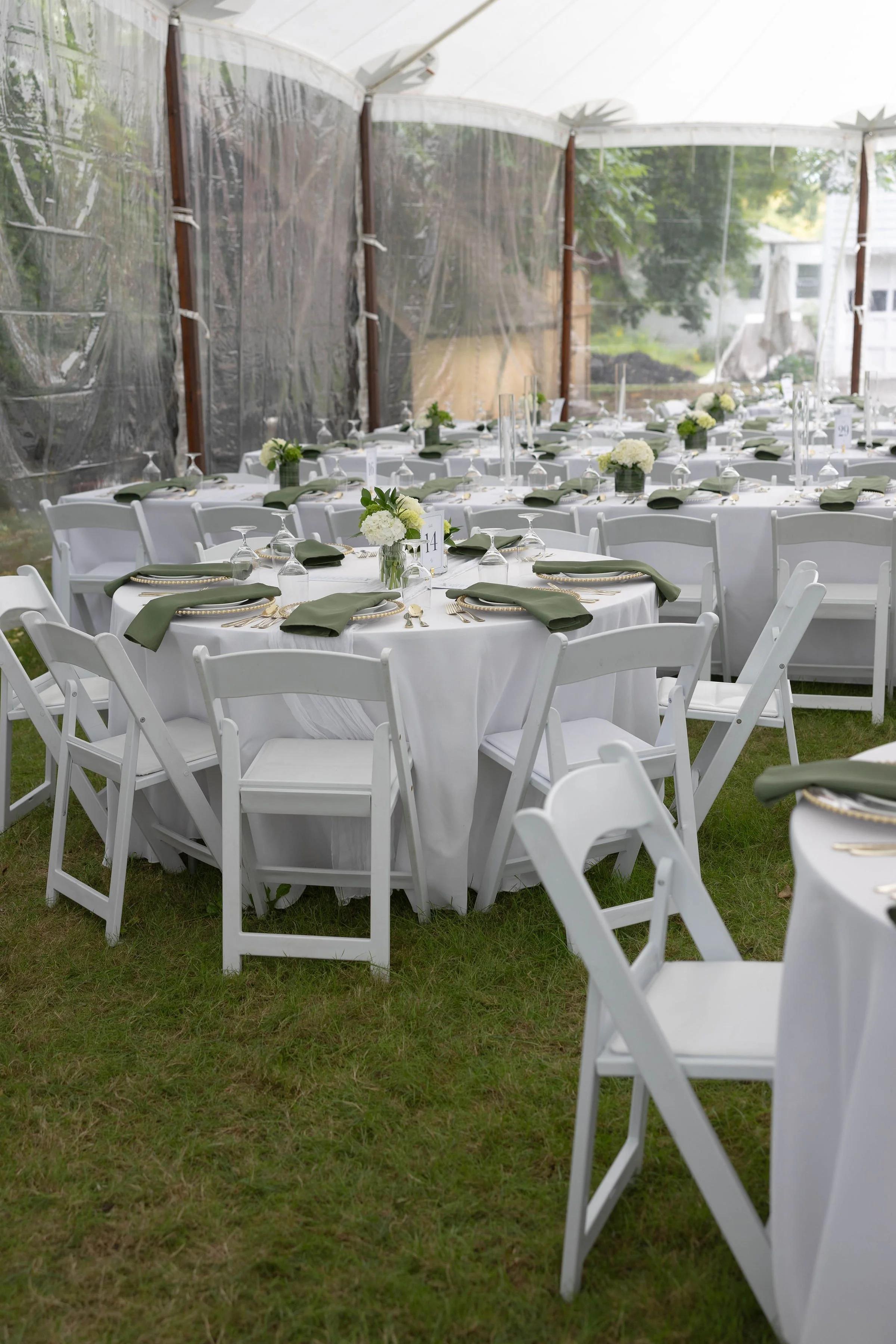 Wedding reception setup with round tables covered in white tablecloths, decorated with white floral centerpieces, green napkins, glassware, and gold utensils, under a white tent with transparent sidewalls.