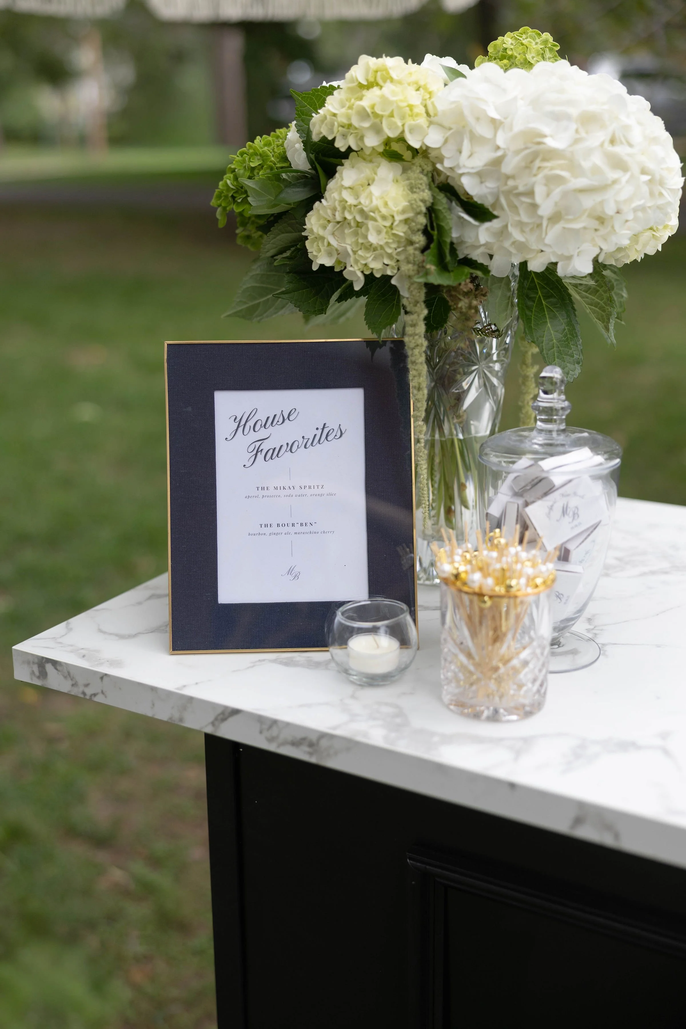A floral arrangement with white hydrangeas and green foliage in a glass vase, a black framed menu card that says 'House Favorites' on a marble table, and small jars with candies or mints, set outdoors with blurred trees and grass in the background.