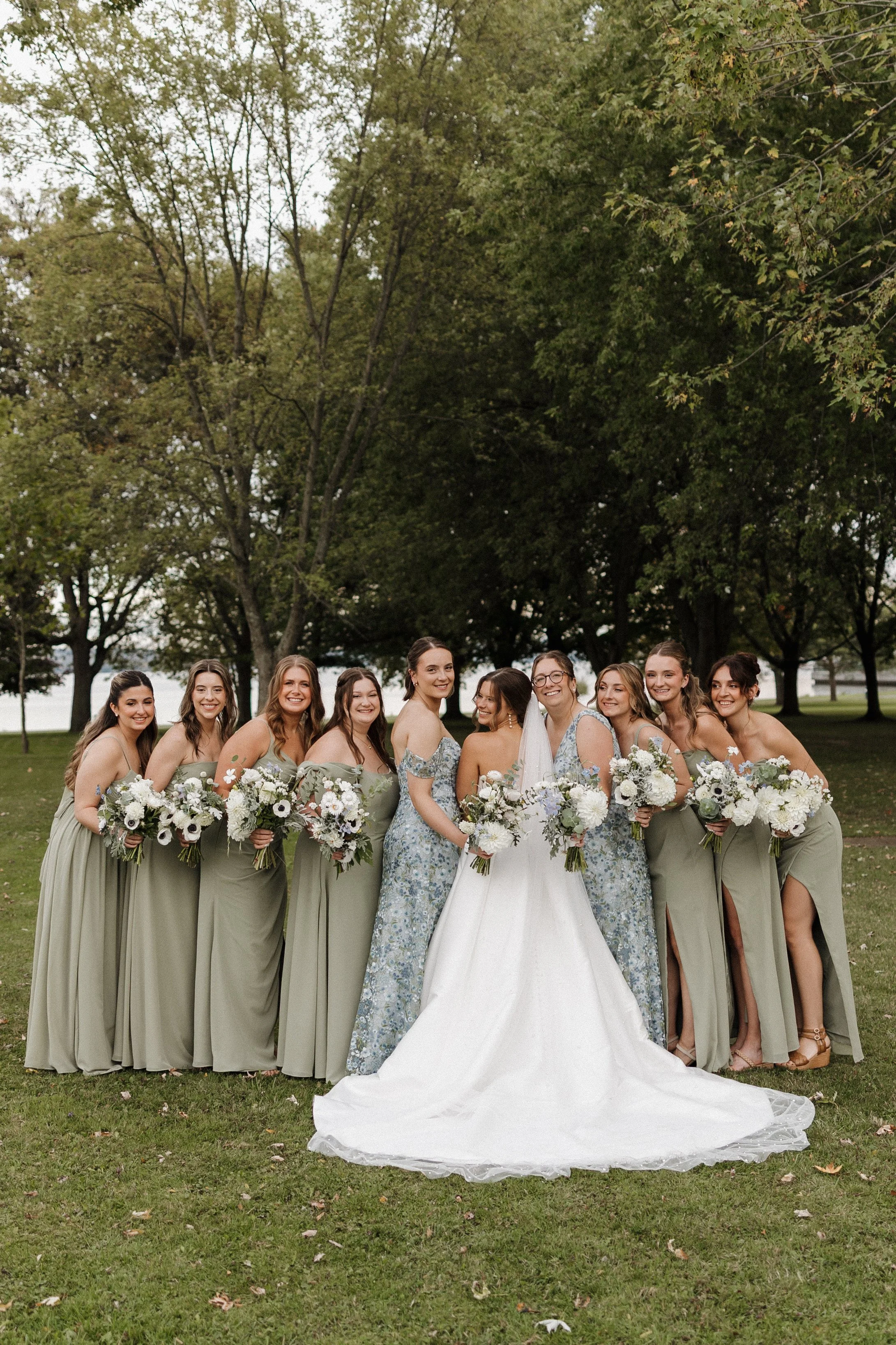 A group of women, including a bride in a white wedding dress, and bridesmaids in light green and floral dresses, standing outdoors on a grassy area with trees in the background, all holding bouquets.