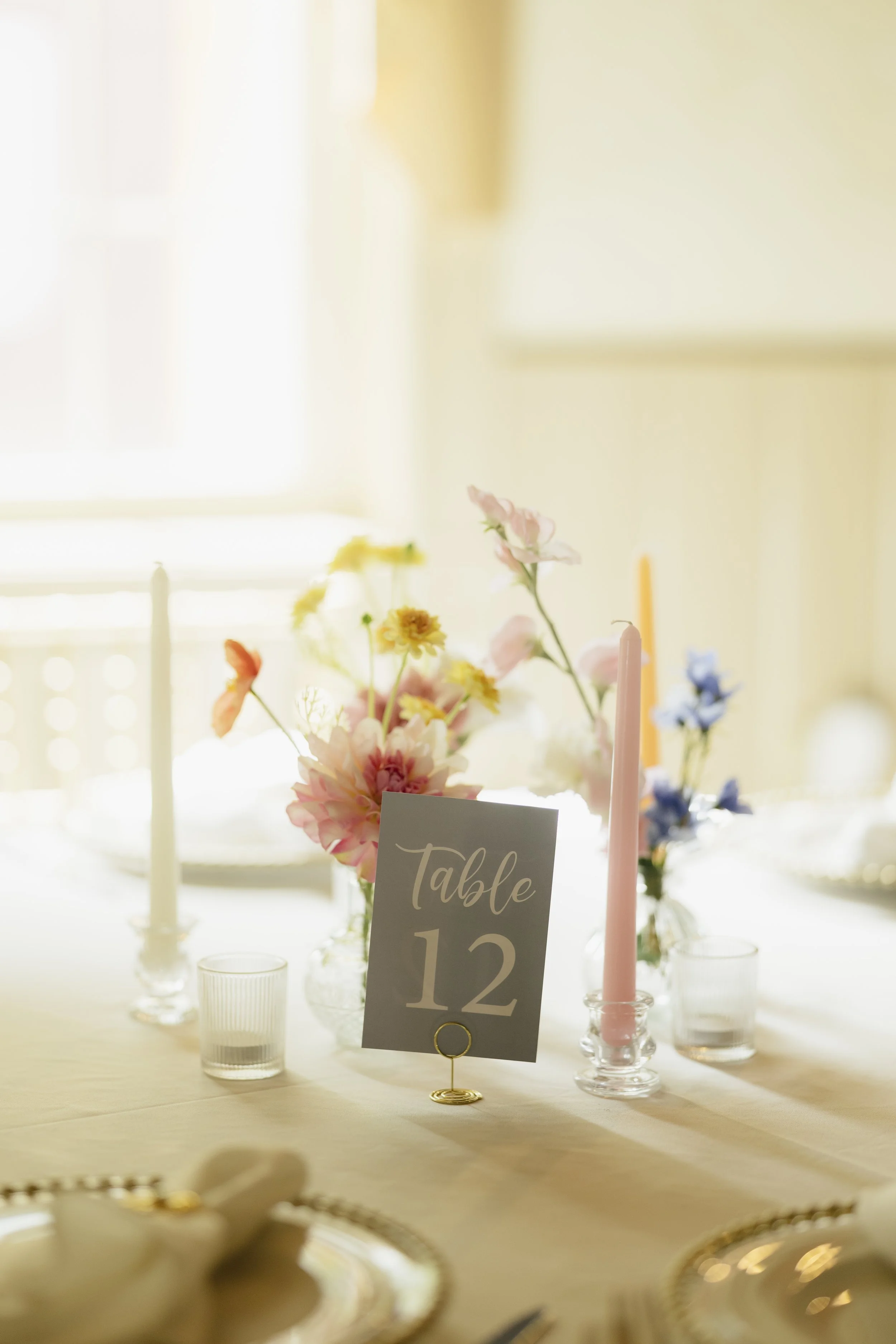 A decorated table setting with a floral centerpiece, pink and pastel candles, and a table number card with the number 12, set in a bright room.