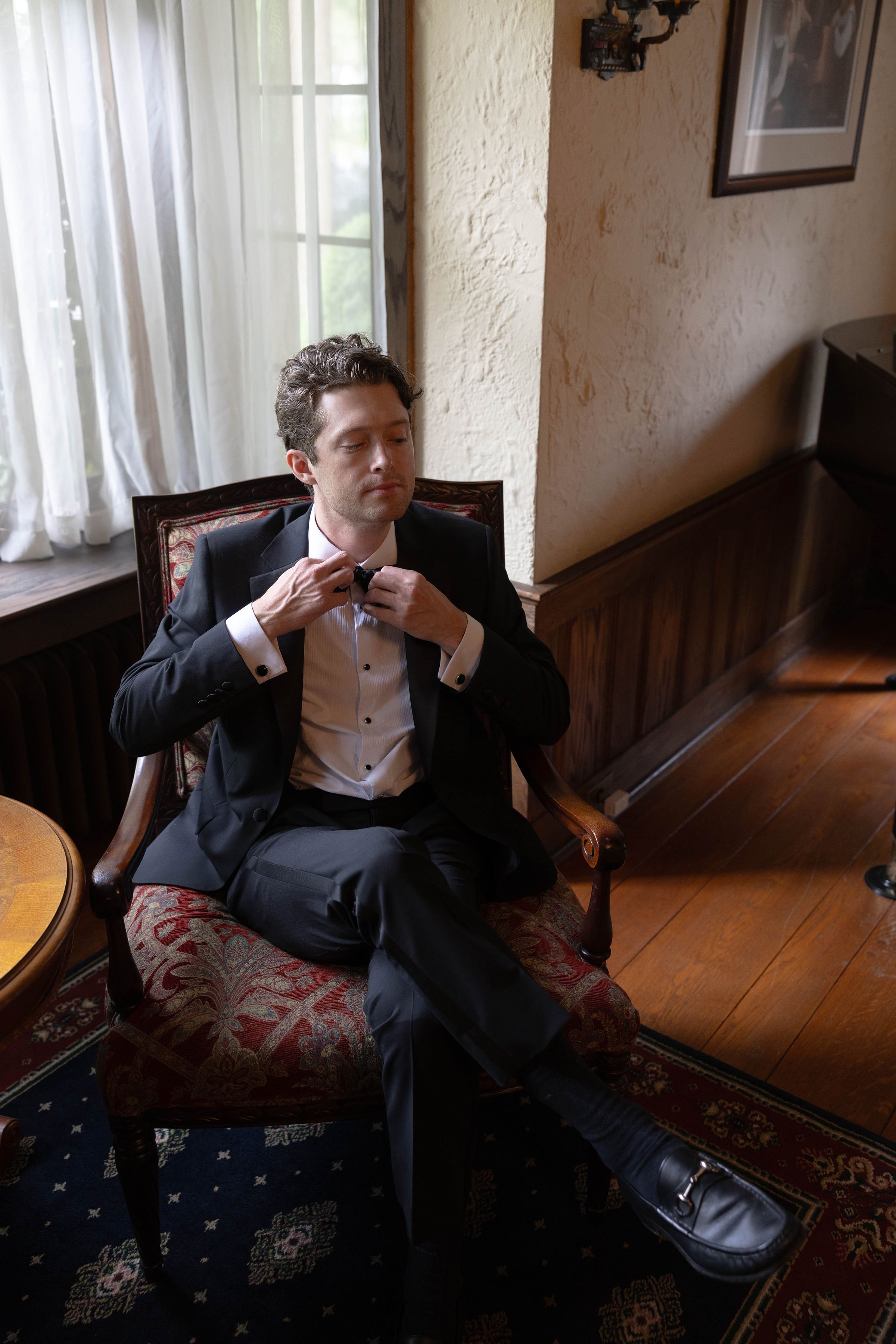 A young man in formal black tuxedo with white shirt and bowtie, sitting on a wooden armchair with patterned red upholstery, adjusting his bowtie. He appears to be waiting or relaxing inside a room with a window and sheer white curtains, a textured be