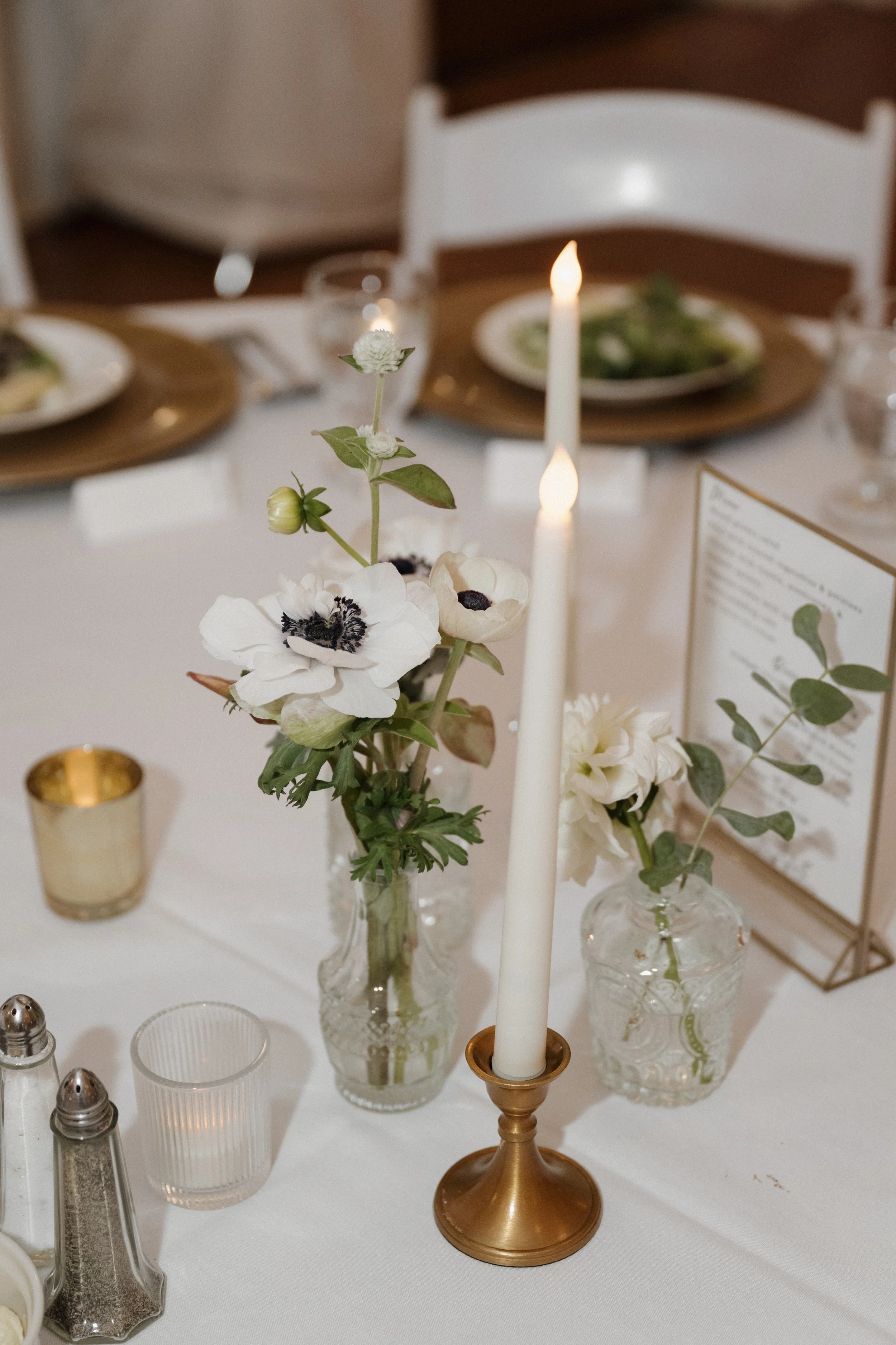 A table decorated with floral arrangements, a lit candle, and various small candles and salt and pepper shakers, set for a formal occasion.