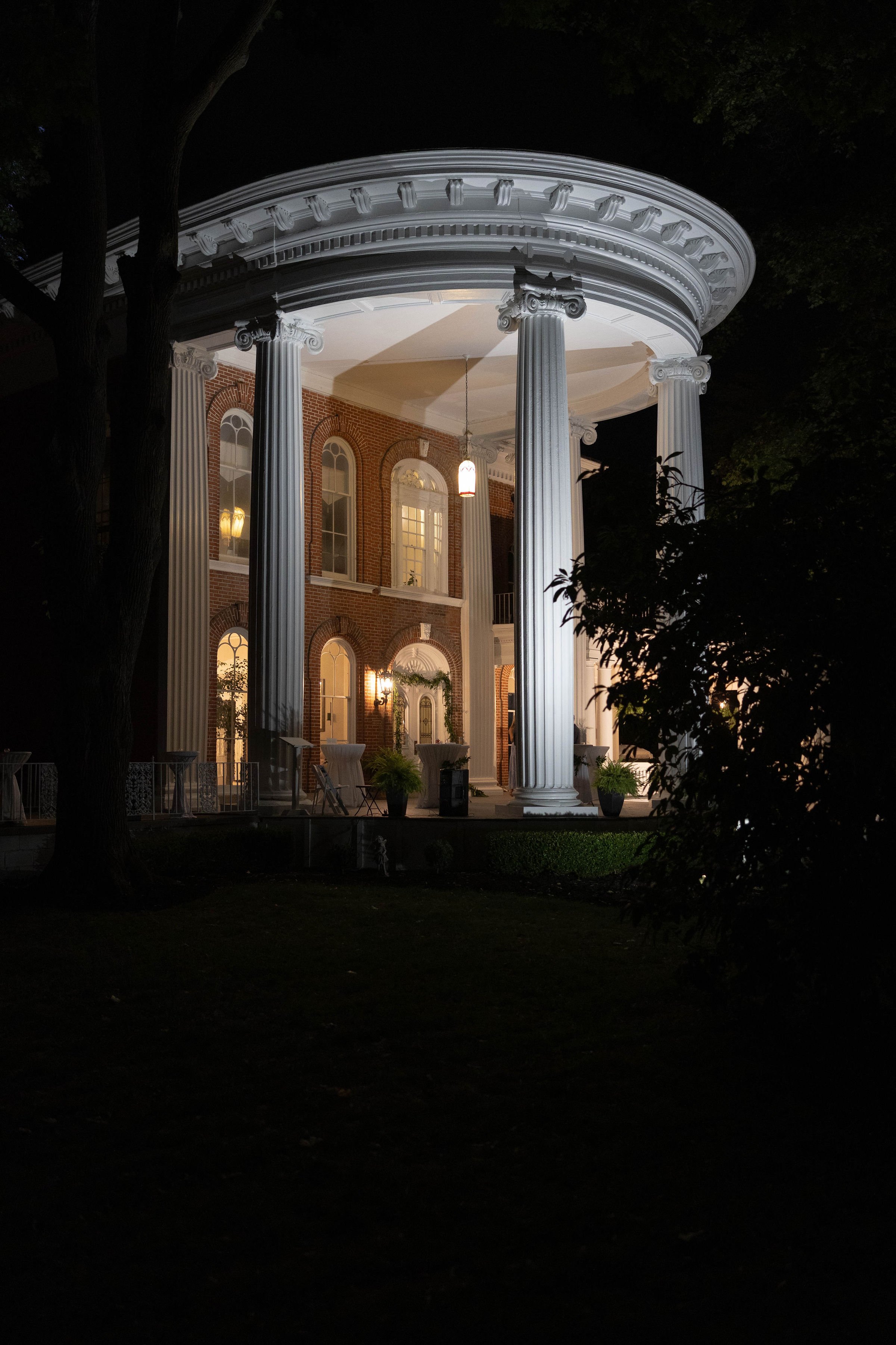 Nighttime view of a large, illuminated house with classical columns and brick walls, partially surrounded by trees.