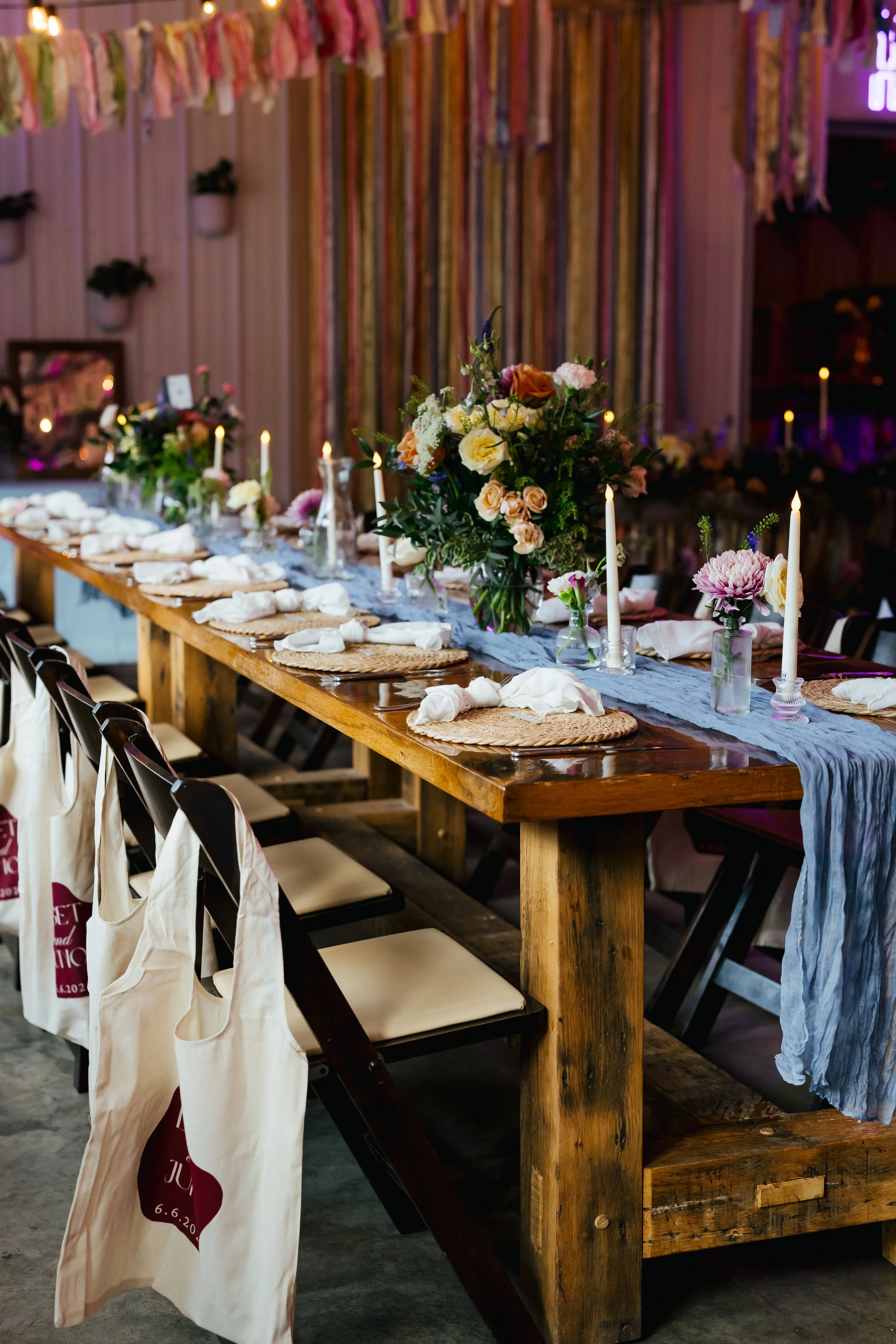 A long wooden banquet table decorated with flower arrangements and candles, set up for a celebration or gathering in a rustic event space with colorful ribbons and greenery on the wall.