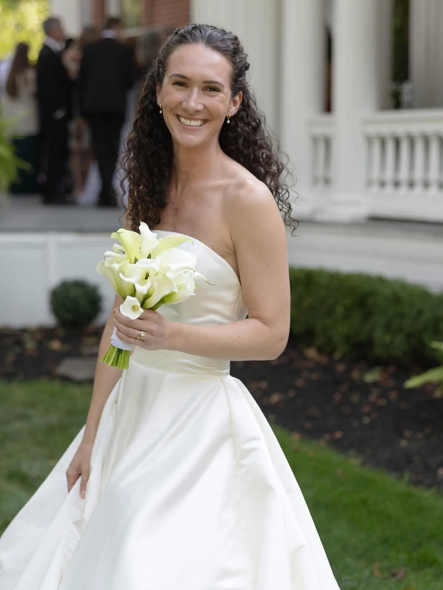 A smiling woman in a wedding dress holding a bouquet of white calla lilies outdoors near a white porch.