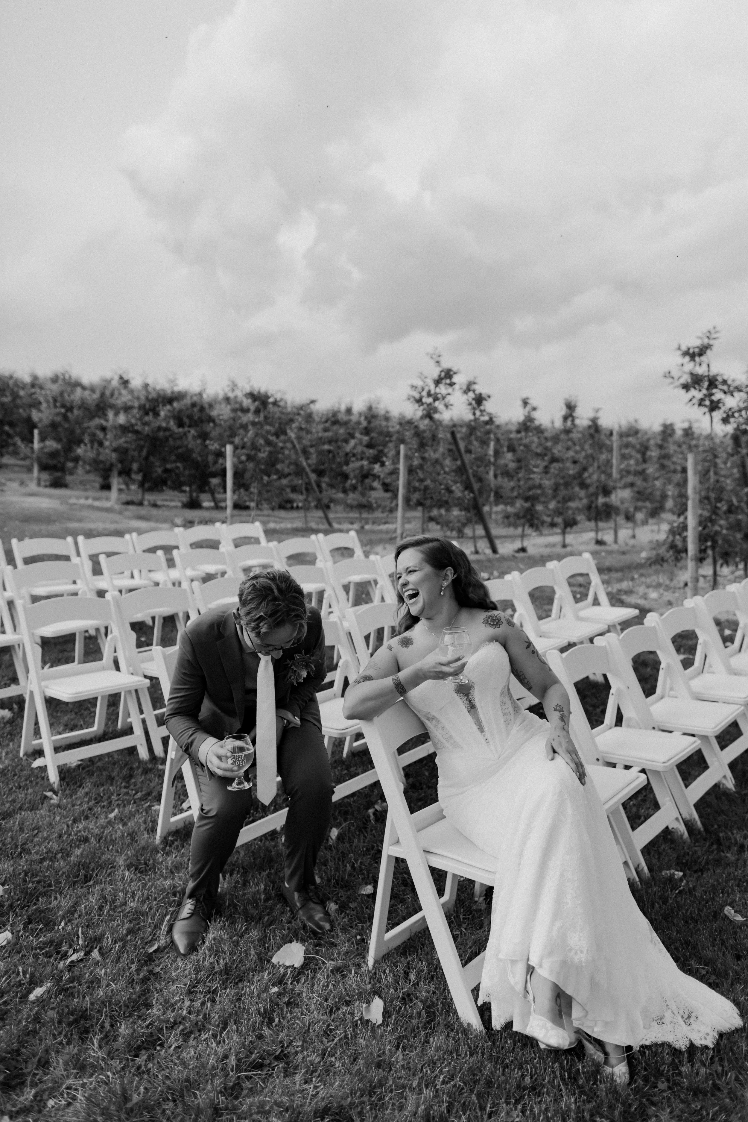 A black-and-white photo of a joyous bride and groom sitting outdoors among empty white chairs at a wedding ceremony. The bride is smiling and holding a wine glass, while the groom appears to be laughing as they lean toward each other. Trees and cloud