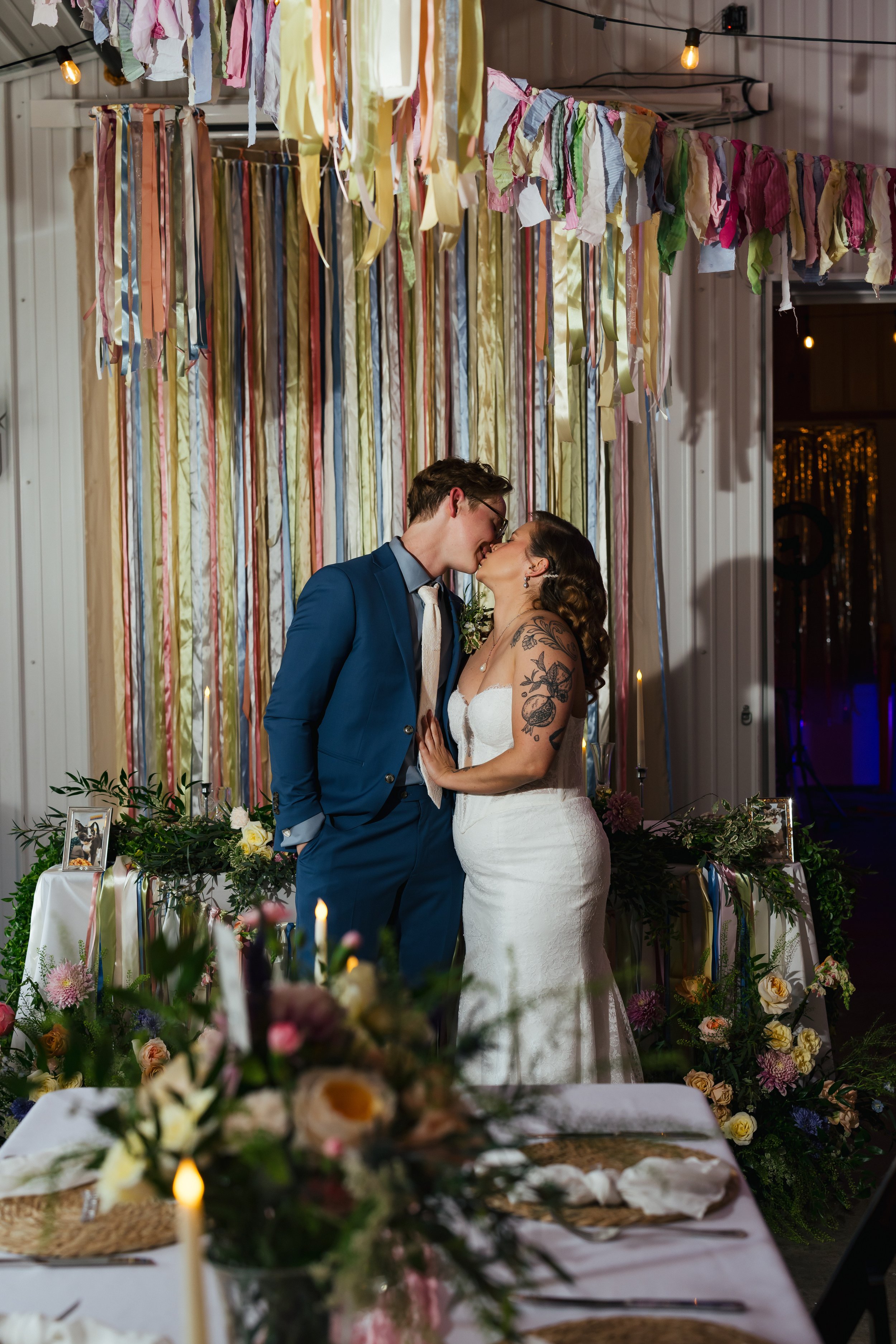 A bride and groom sharing a kiss at their wedding reception. The bride has tattoos on her arm and is wearing a white wedding dress. The groom is dressed in a blue suit with a white tie. There are colorful ribbons hanging from the ceiling, and a decor