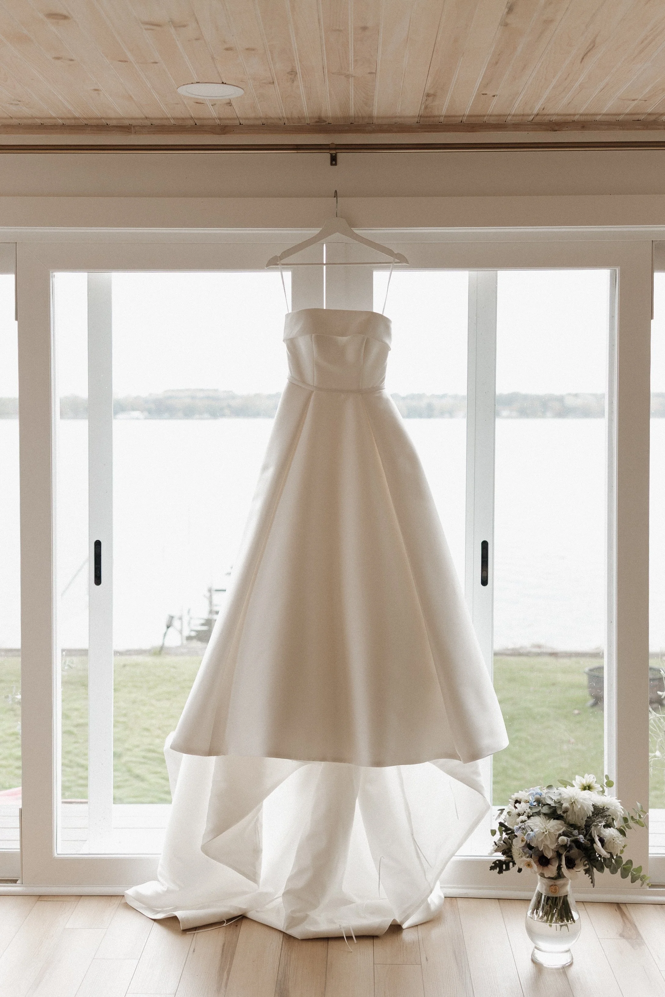 A wedding dress hanging on a hanger in front of a large window with a view of a body of water and trees outside. A bouquet of flowers is placed in a glass vase on the floor.