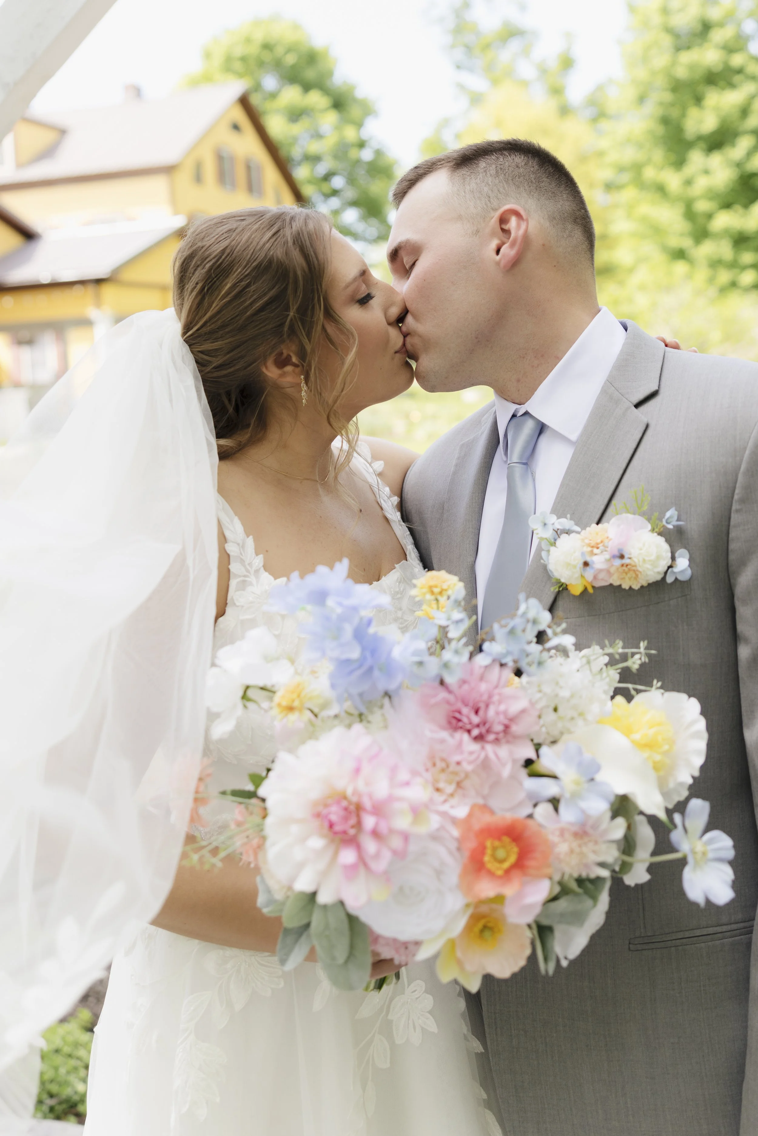 A bride and groom share a kiss outdoors on a bright day. The bride wears a white wedding dress and veil, and the groom wears a light gray suit with a boutonniere. The bride holds a large bouquet of colorful flowers, and there are trees and a yellow h