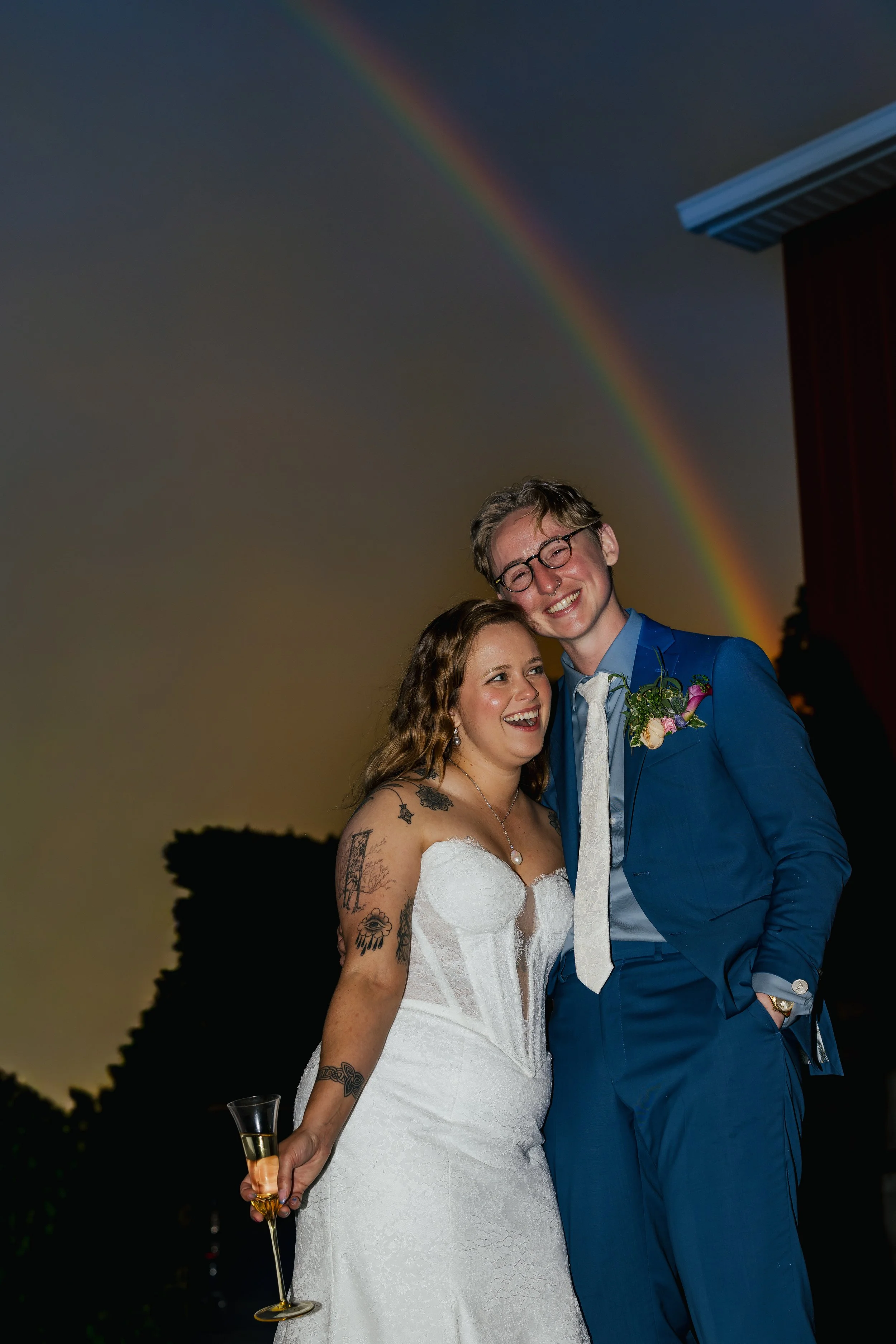 A happy couple at their wedding, standing close together outdoors at sunset, holding a champagne flute, with a rainbow in the sky behind them.