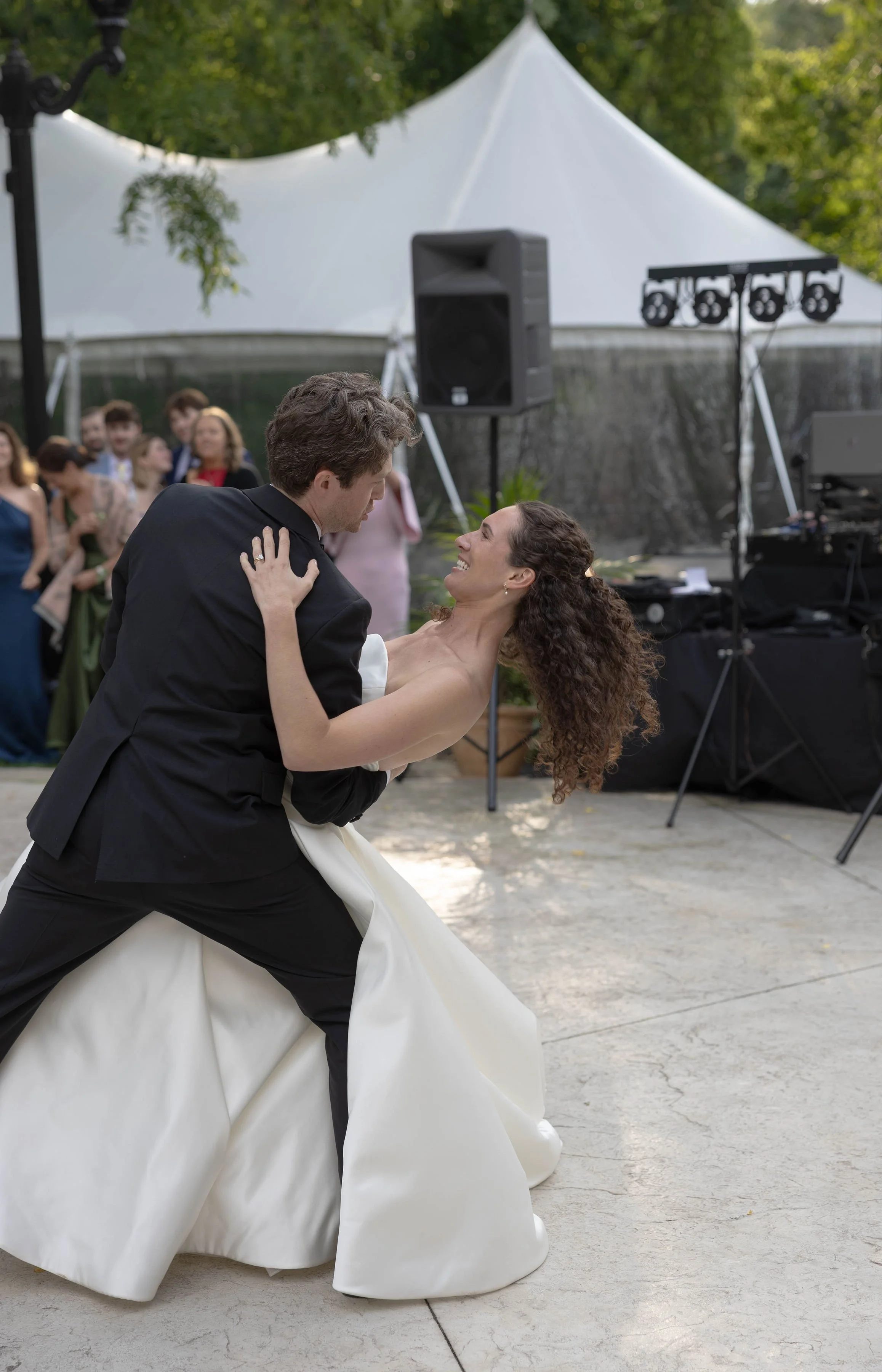 A bride and groom dancing at an outdoor wedding reception, with guests and a DJ setup in the background.