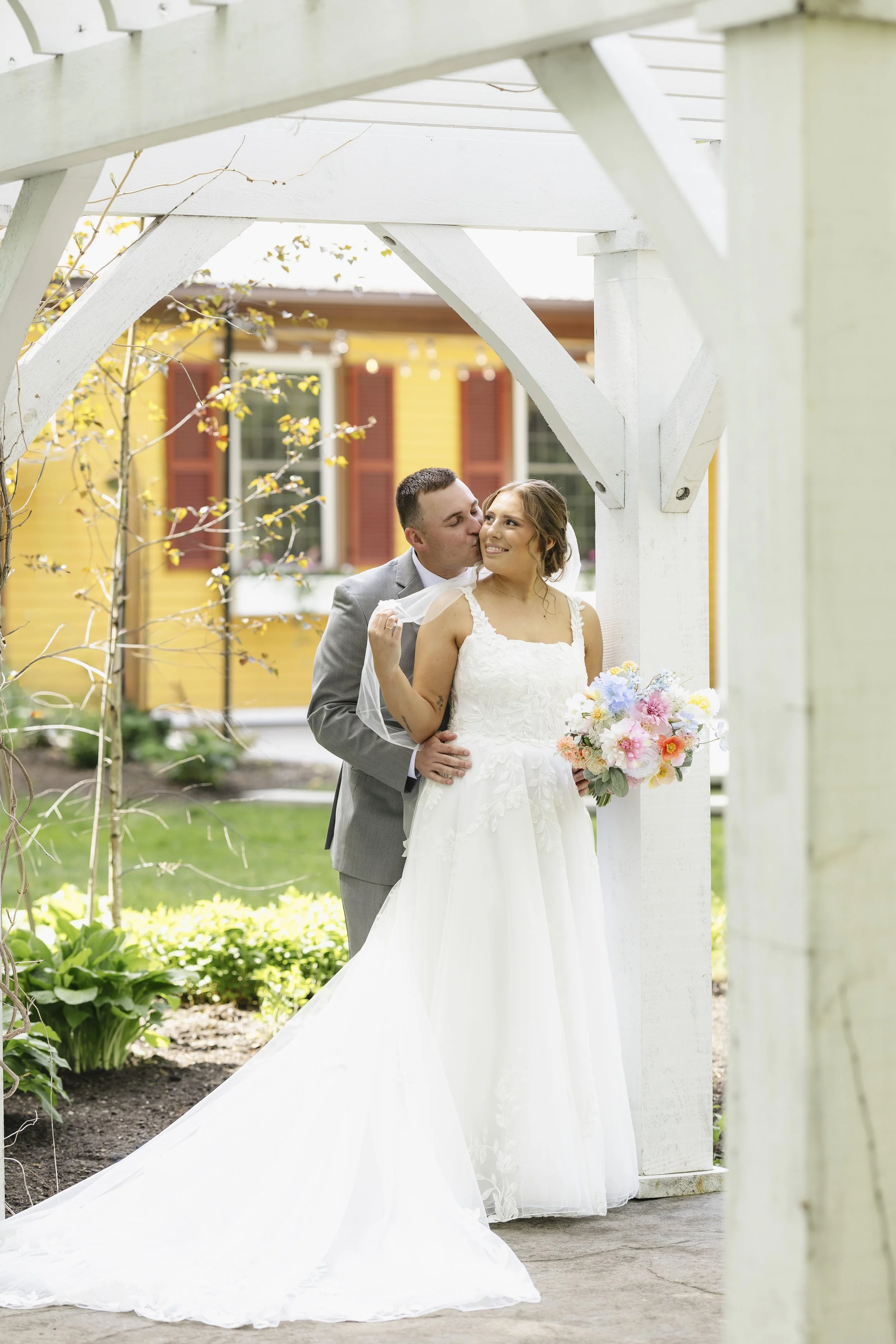 A bride and groom at their wedding, standing outdoors under a white wooden arbor. The bride is smiling and holding a bouquet of pink, white, and purple flowers, while the groom is kissing her cheek from behind. The background features a yellow house 