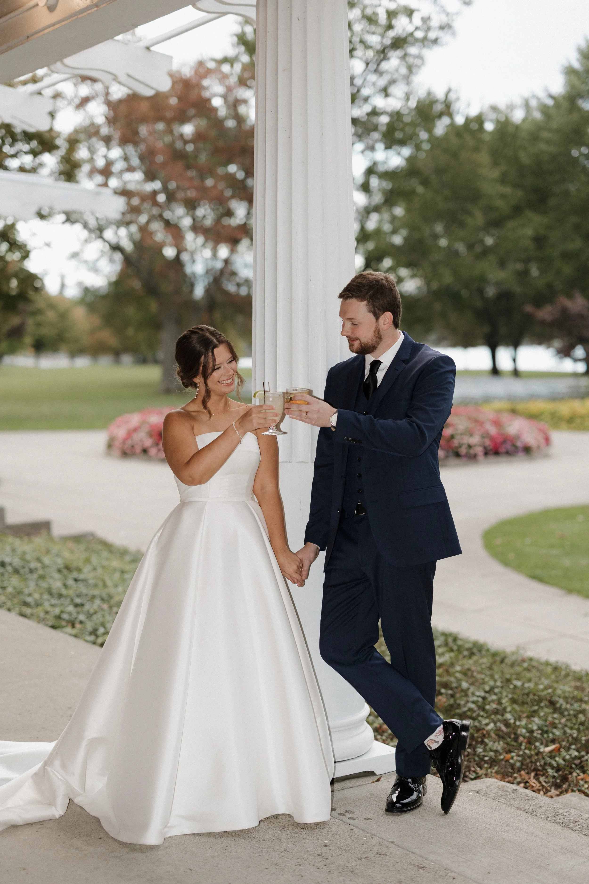 A bride and groom standing outdoors near a white column, holding glasses and toasting each other, with trees and a flower bed in the background.