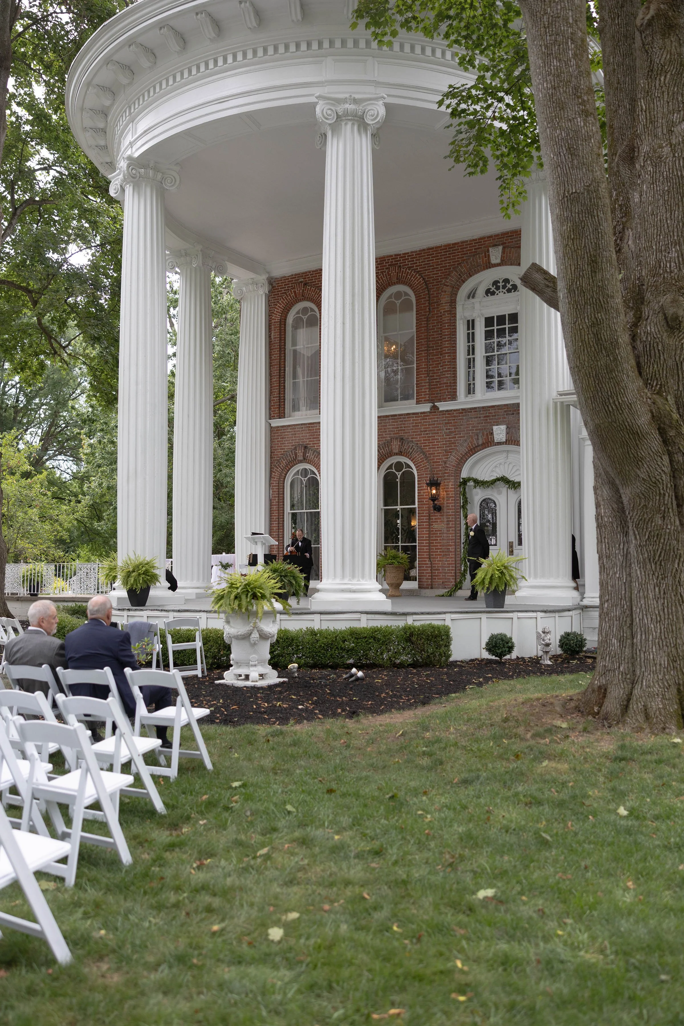 A wedding ceremony setup outdoors in front of a large, historic brick building with white columns and a portico. White chairs are arranged on the grass for guests, and a small group of people is gathered near the entrance. The scene includes trees an
