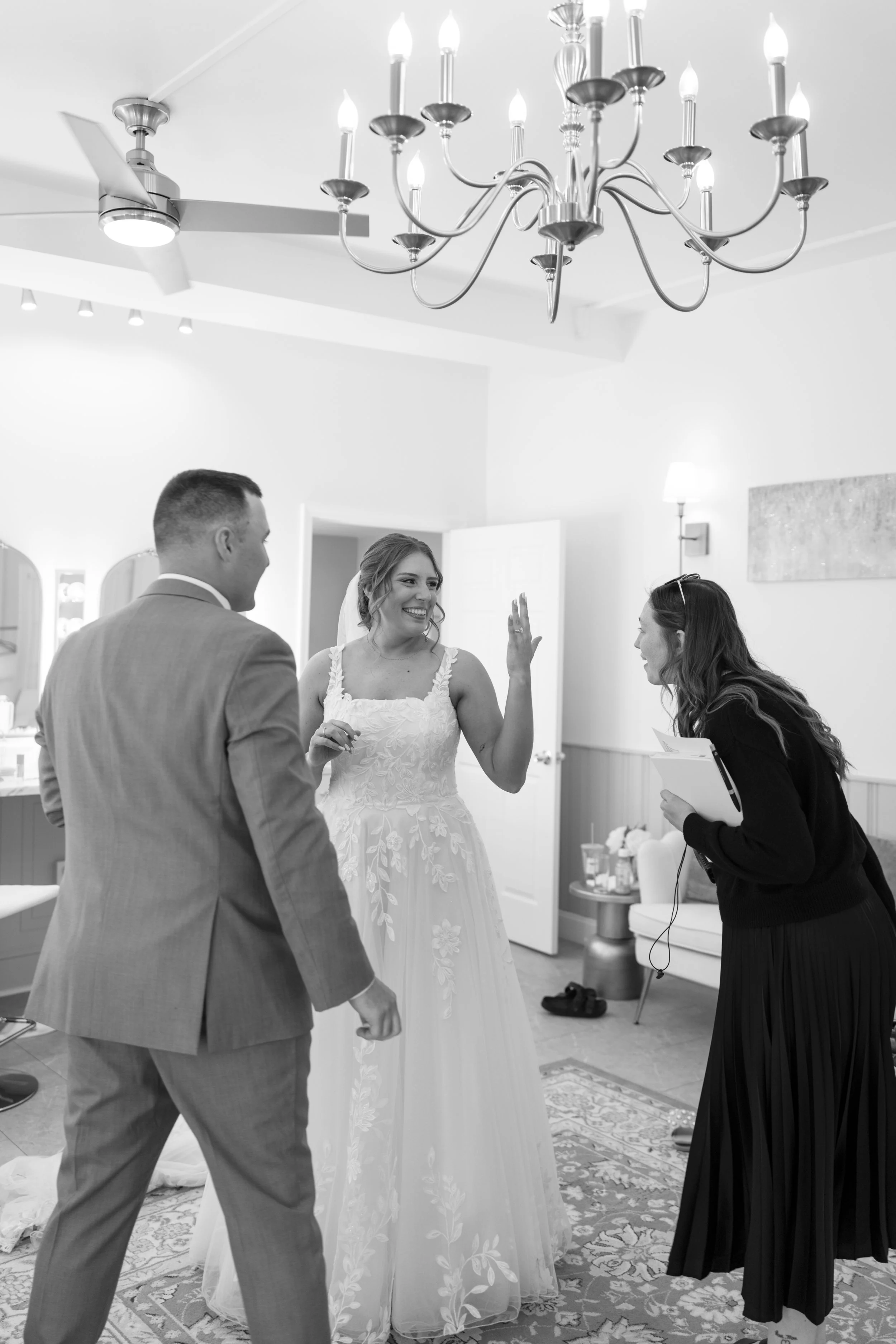 A wedding ceremony with a bride in a floral lace gown and veil, a groom in a suit, and a woman officiant speaking to the bride. The room has a chandelier, ceiling fan, and decorative rugs.
