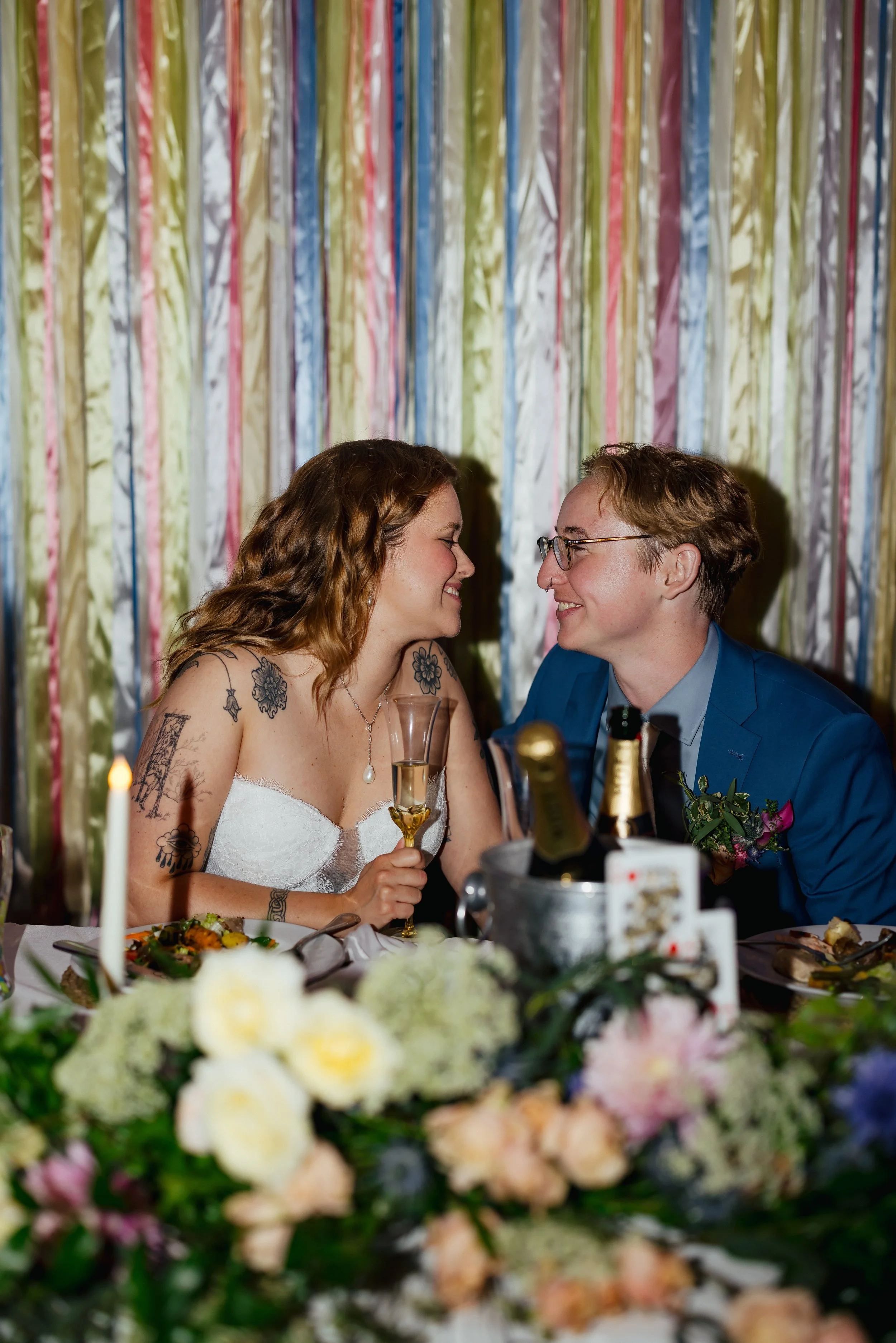 A couple smiling and looking at each other at a wedding reception, seated at a table with food, drinks, and a floral centerpiece, with colorful streamers in the background.