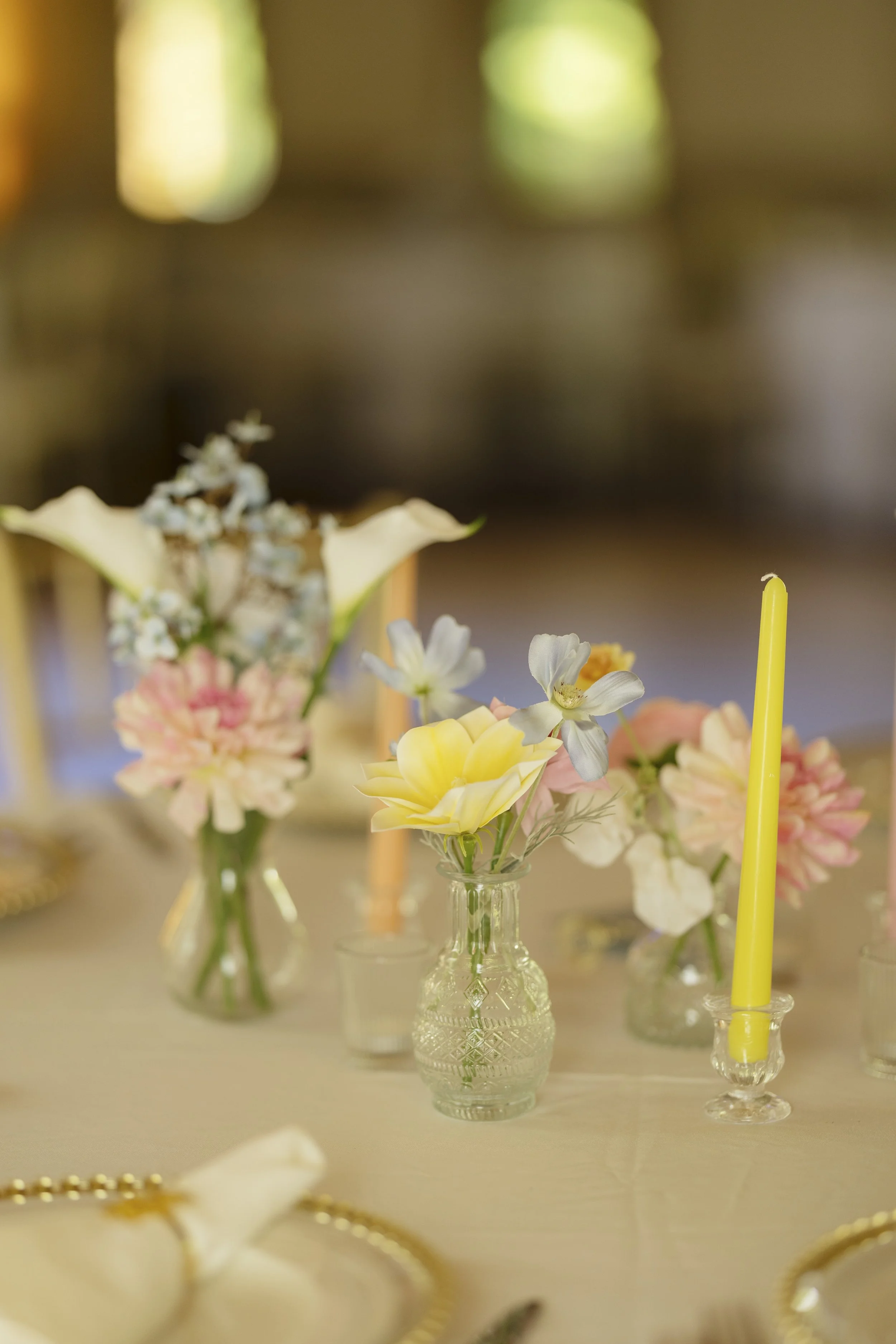 Multiple small flower arrangements in glass vases with white and pink flowers, and yellow candles on a beige table, likely for a celebration or event.