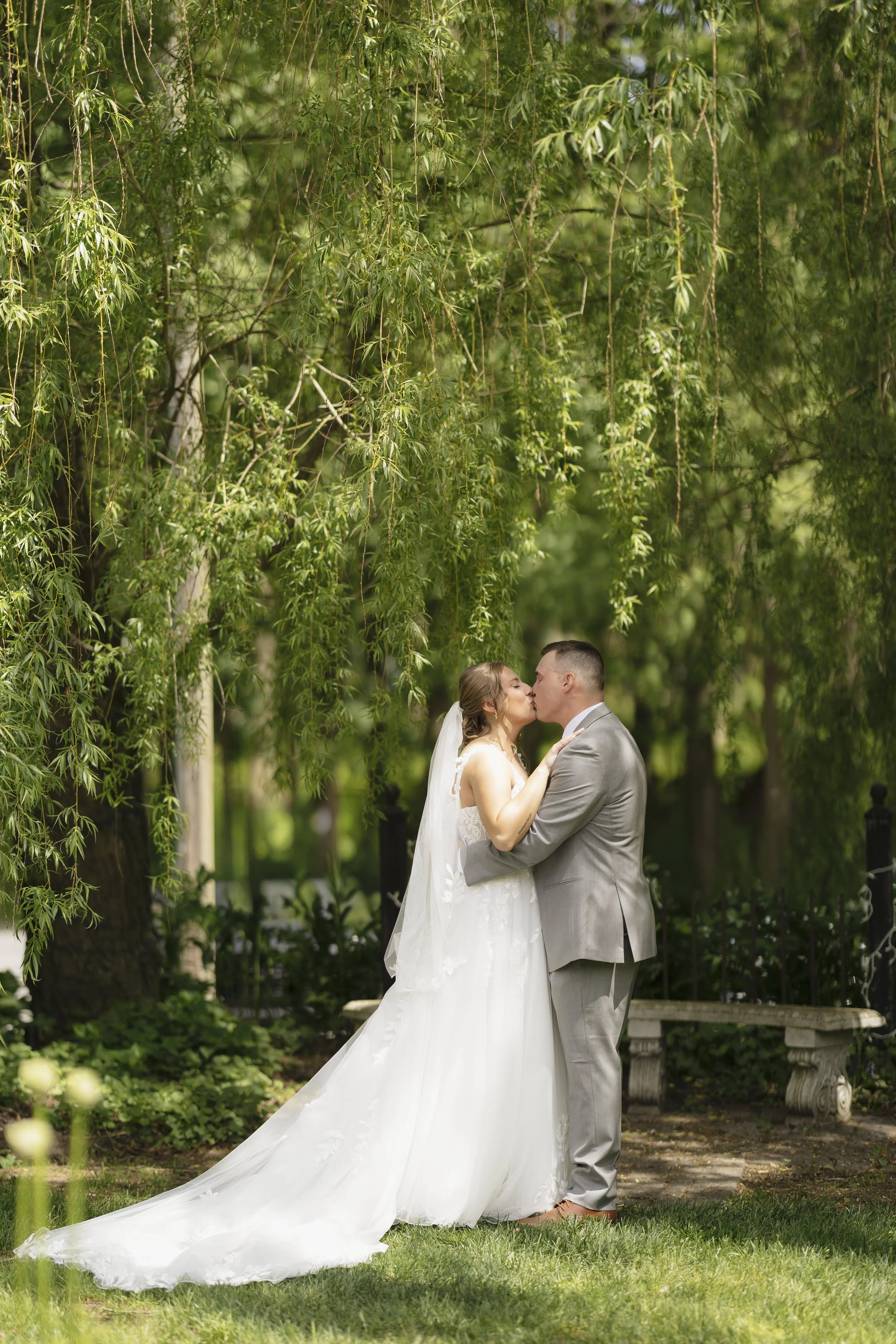 A bride and groom kiss in a green outdoor park under hanging tree branches, with the bride in a white wedding gown and the groom in a light gray suit.