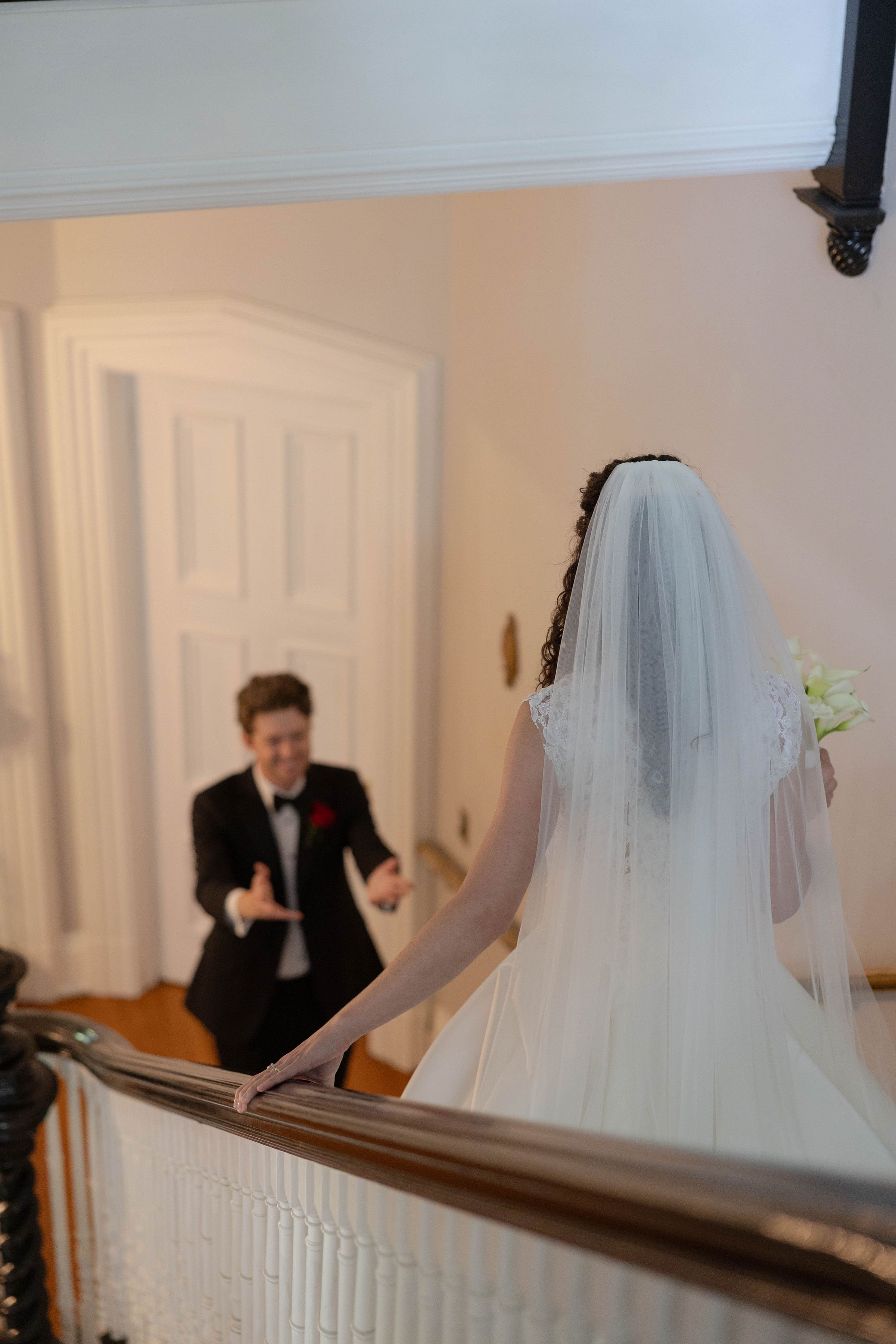 A bride in a wedding dress and veil standing on a staircase, looking at a young boy in a tuxedo smiling and gesturing towards her.