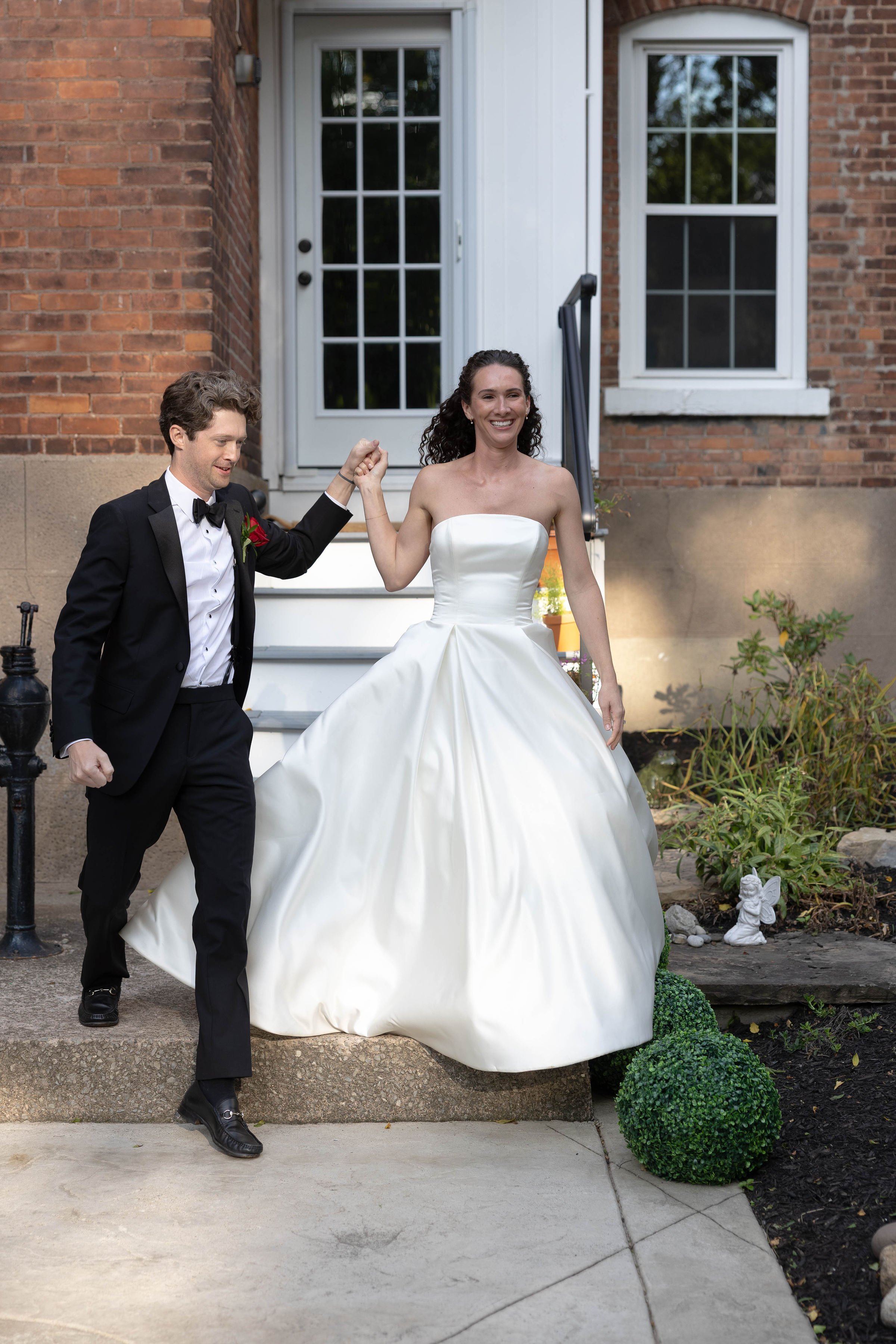 A bride and groom are standing outside a brick house, holding hands and smiling. The bride is wearing a white strapless wedding gown, and the groom is dressed in a black tuxedo with a bow tie.