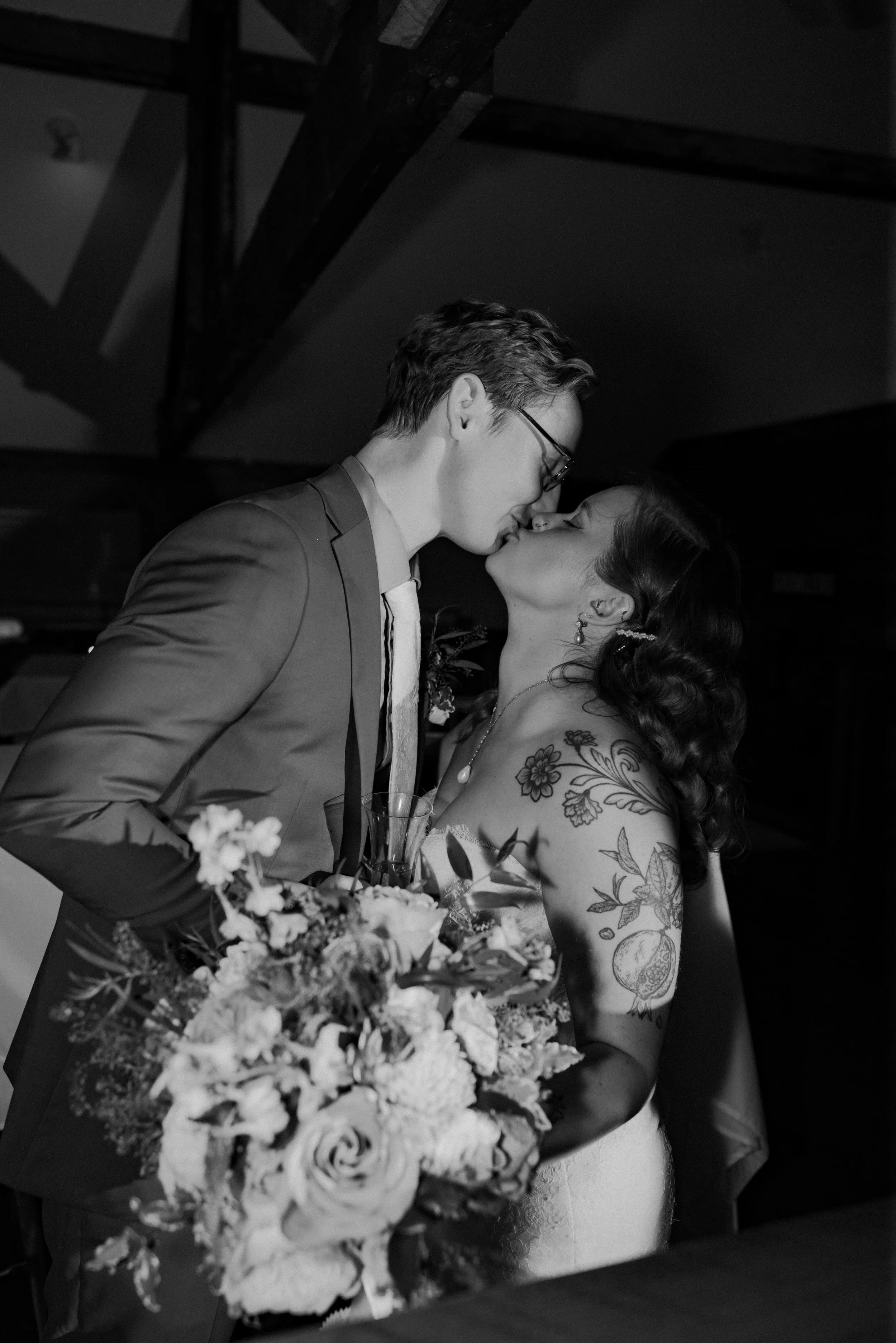 A black and white photo of a couple kissing at a wedding reception. The woman has floral tattoos on her arms and is holding a bouquet, while the man is wearing a suit and glasses.
