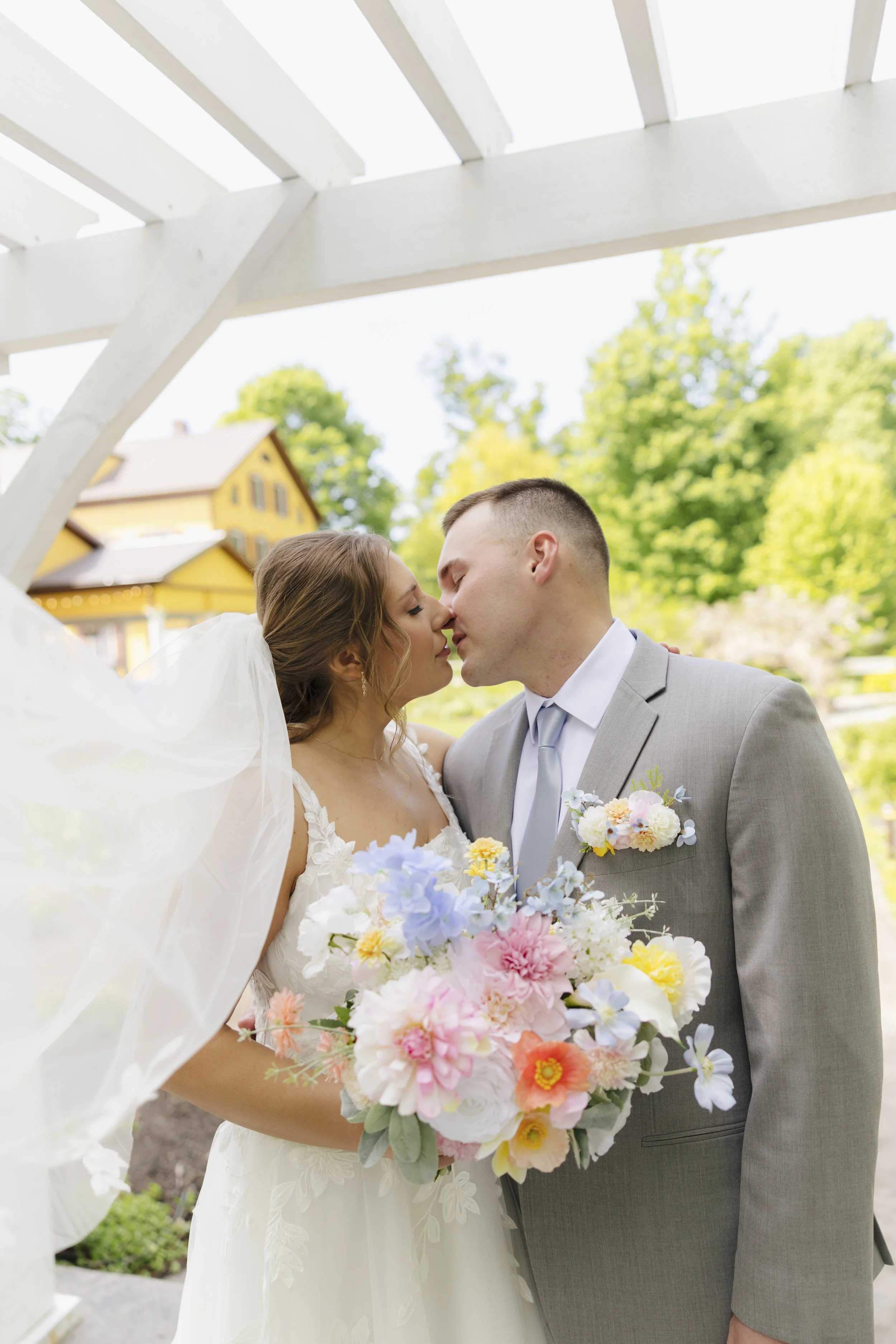 A bride and groom sharing a kiss during their outdoor wedding ceremony, with the bride holding a large bouquet of colorful flowers, and a yellow house and green trees in the background.