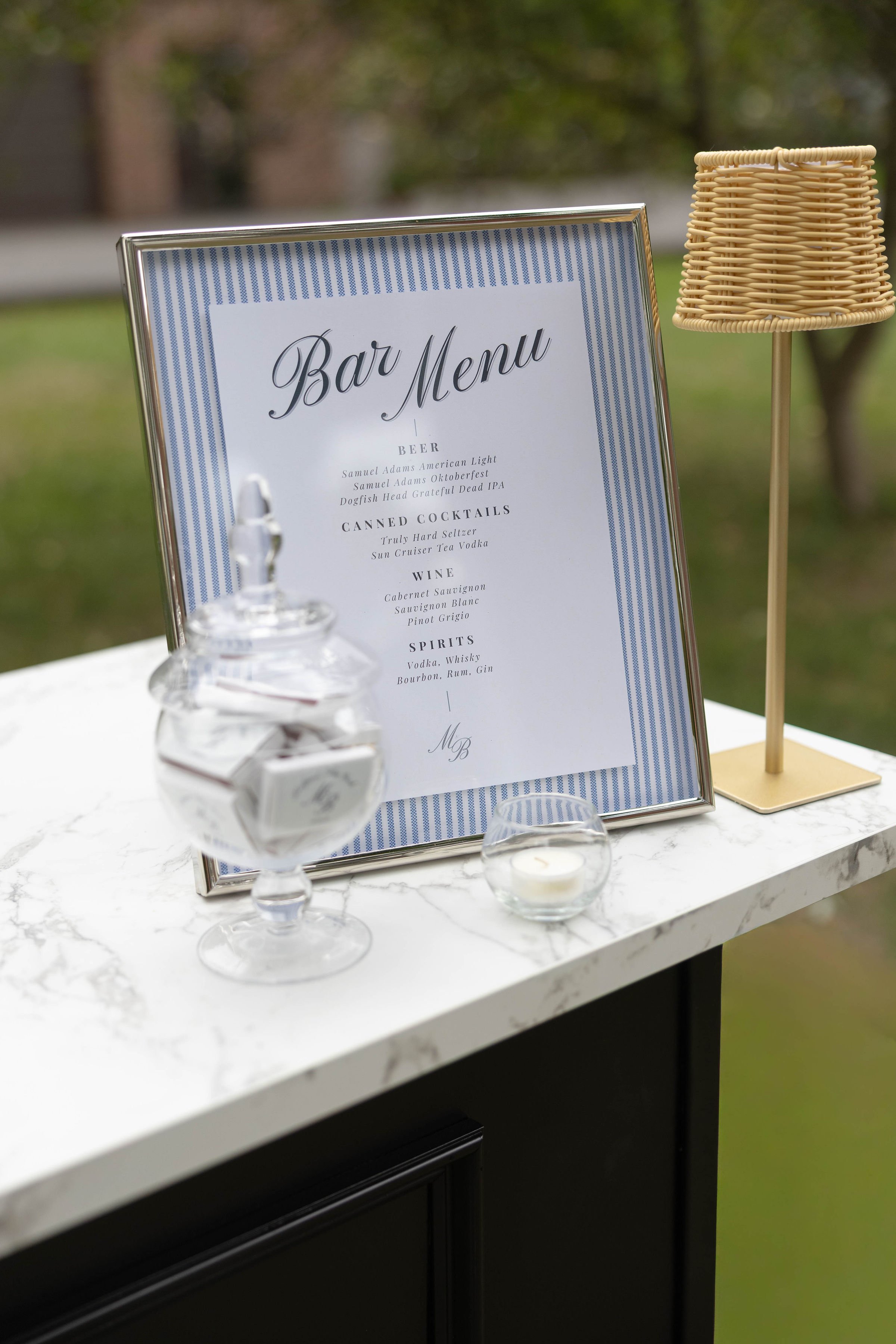 A framed bar menu on a white marble table, with a small glass container and a white candle in front, and a wicker lamp to the right. The background is blurred with outdoor scenery.