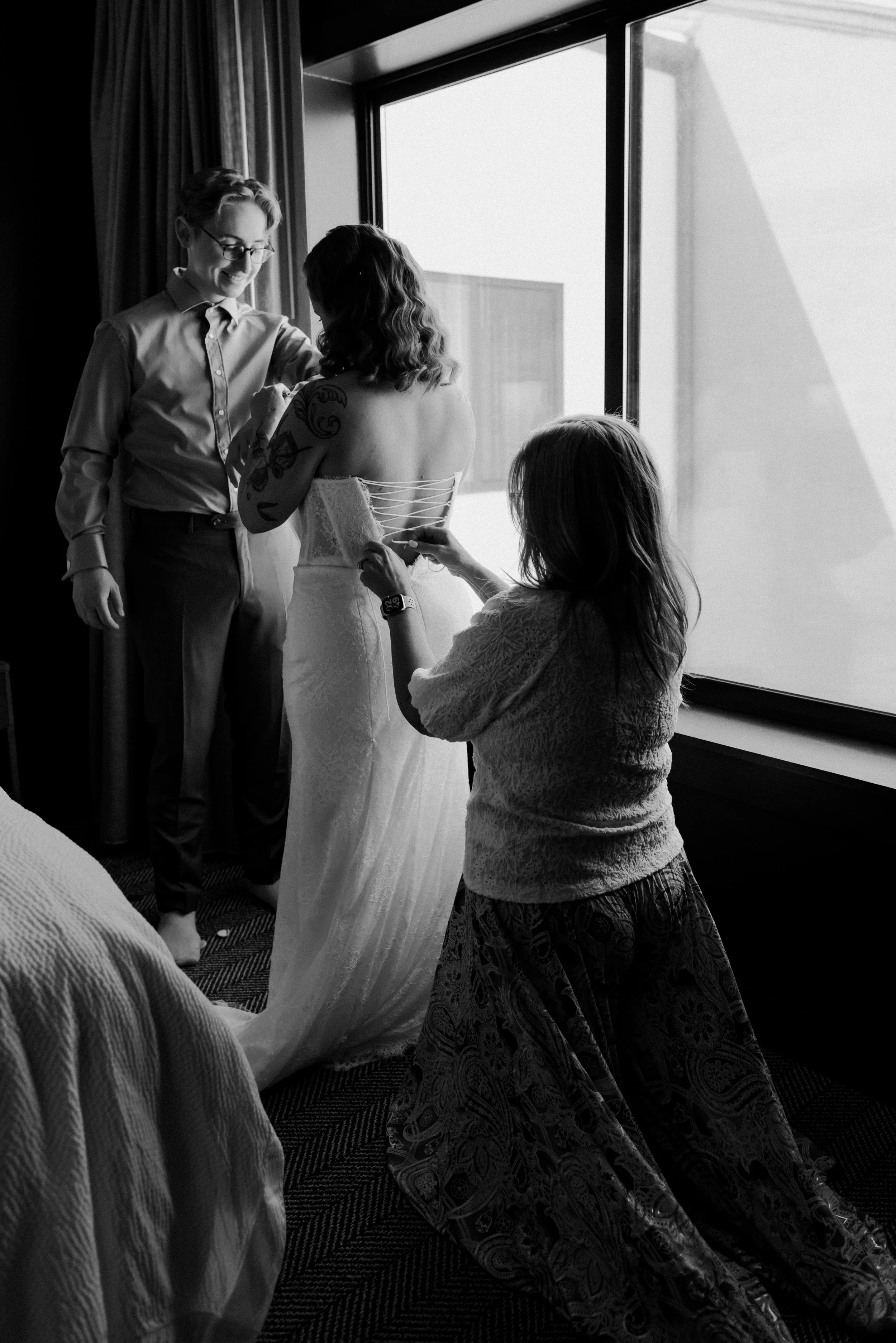 A woman in a wedding dress is getting ready, assisted by another woman while a young girl watches. They are near a large window with natural light streaming in.