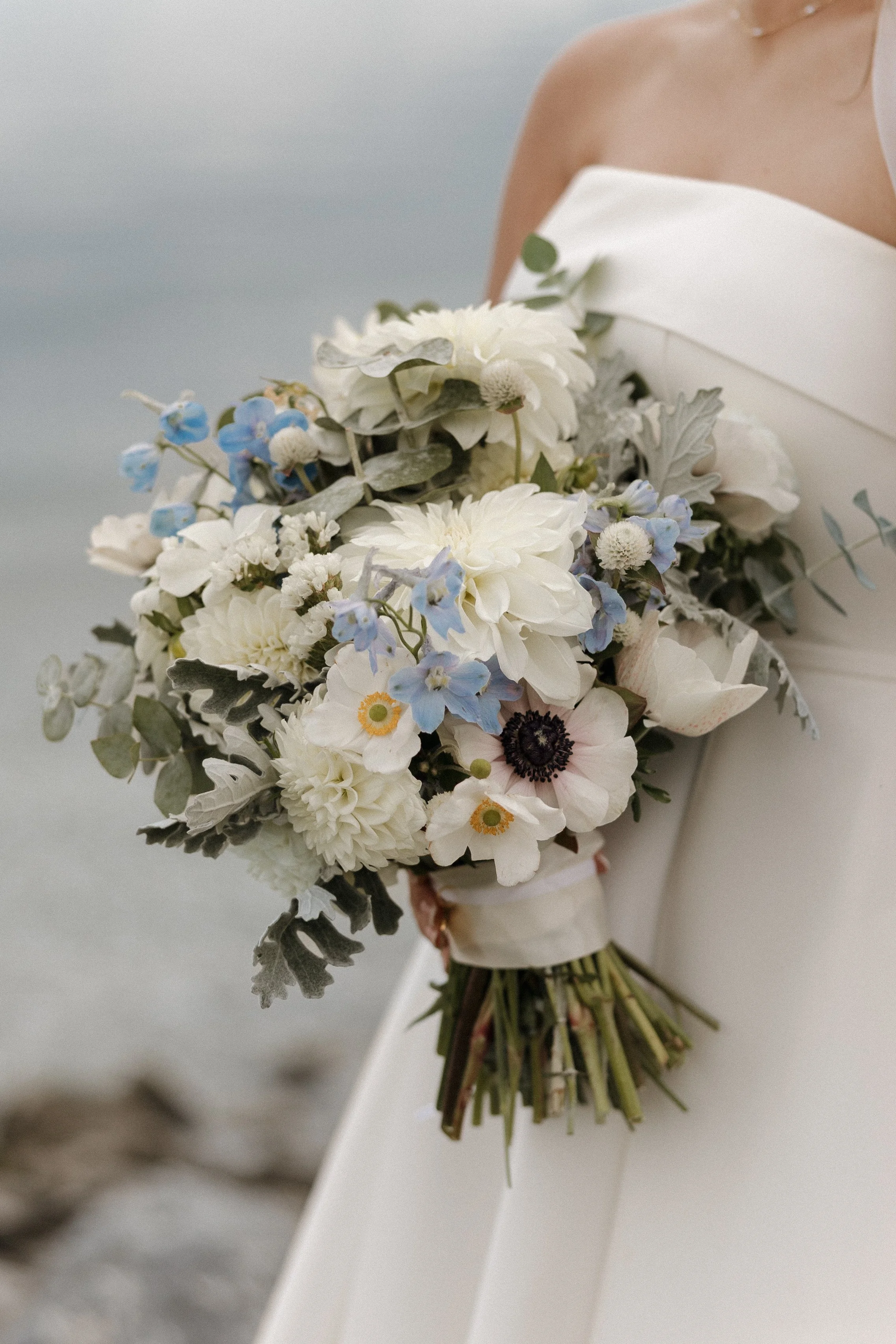 A bride in a strapless white wedding dress holding a bouquet of white, blue, and green flowers.