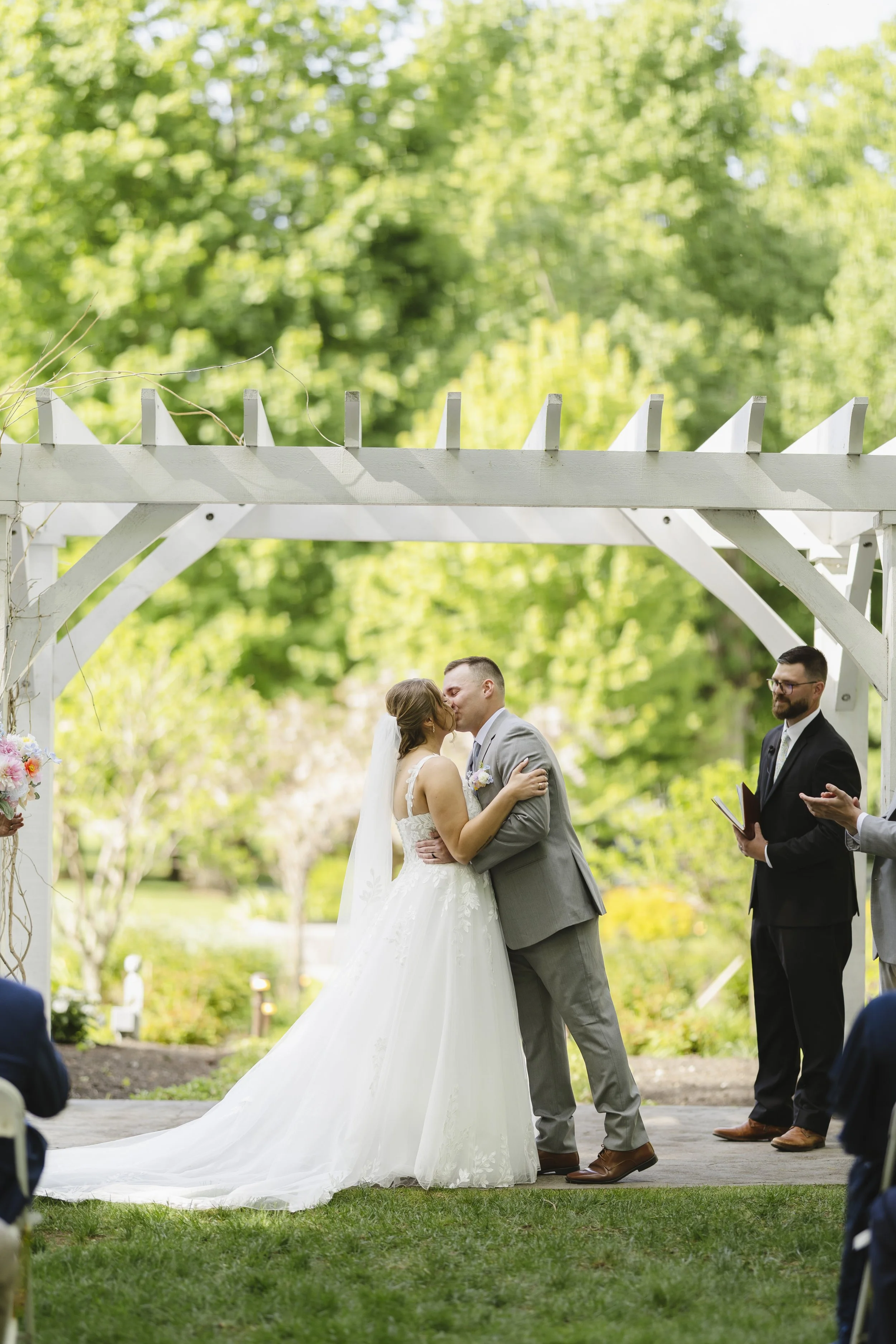 A bride and groom share a kiss at their outdoor wedding ceremony, with a officiant nearby and guests watching, set in a lush green garden with a white wooden arch.
