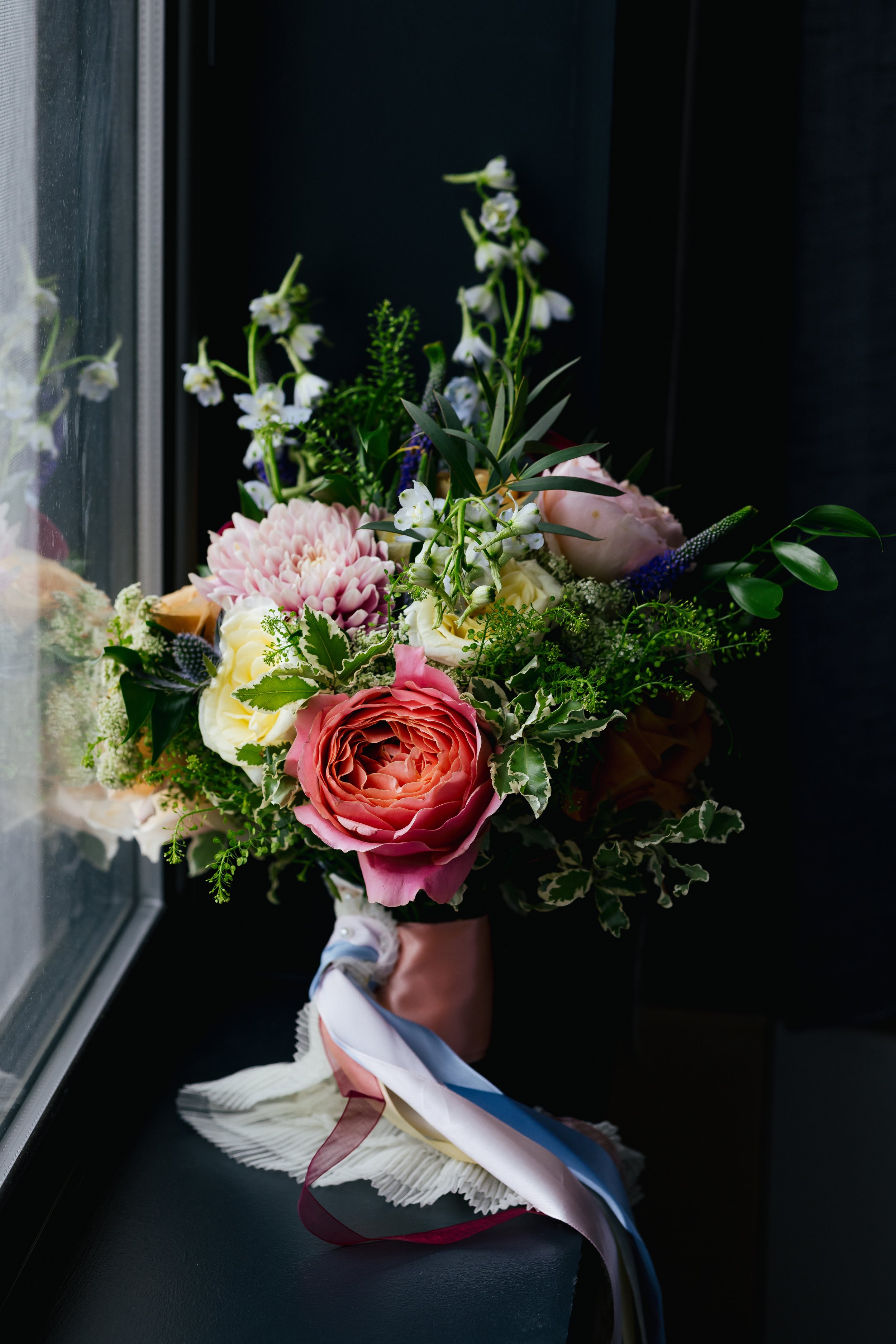 A flower bouquet held by a hand with a decorative ribbon, placed on a black surface near a window with raindrops on the glass.