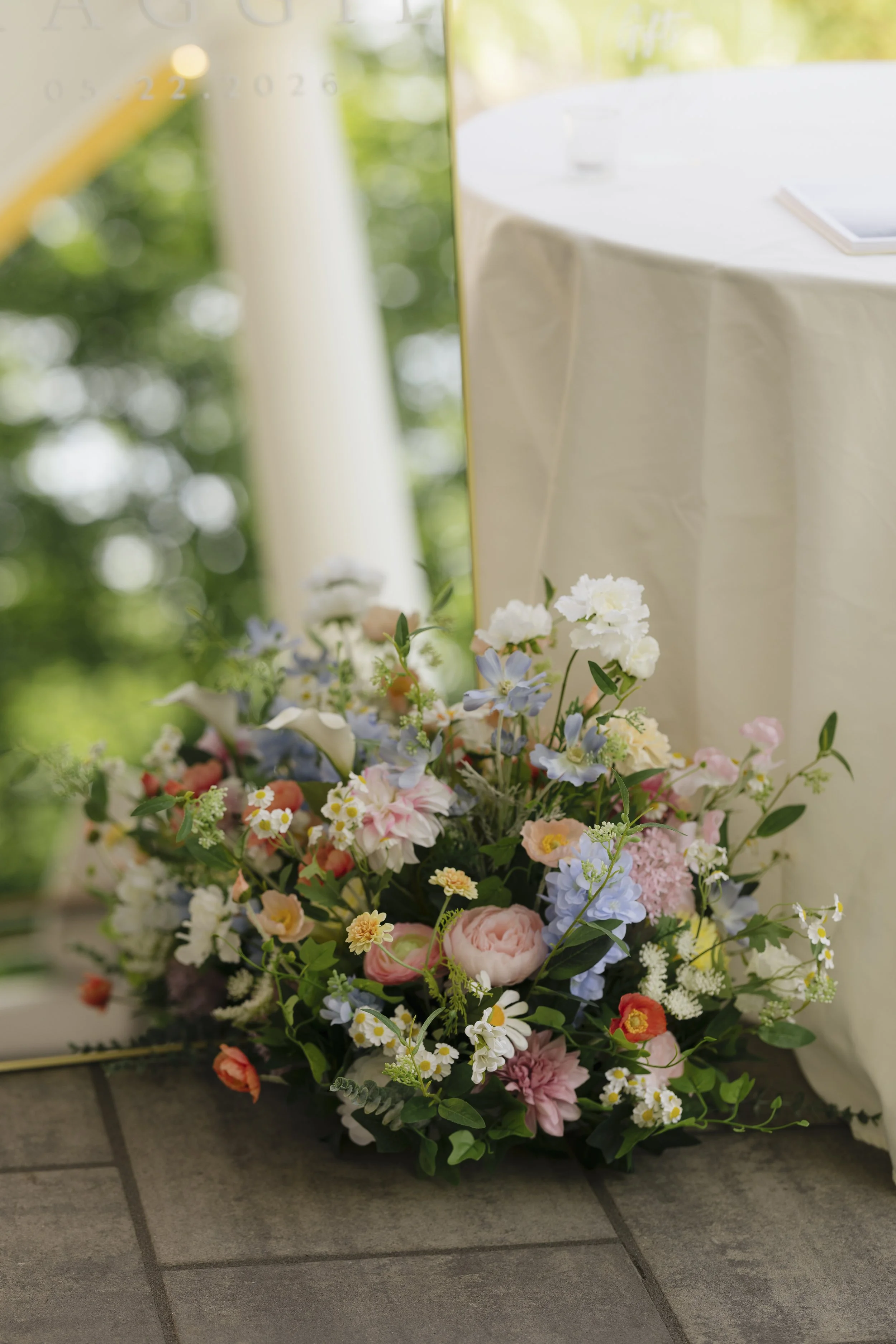 A colorful floral arrangement with pink, white, purple, and yellow flowers placed on a tiled floor in front of a blurred background and a table with a white cloth.