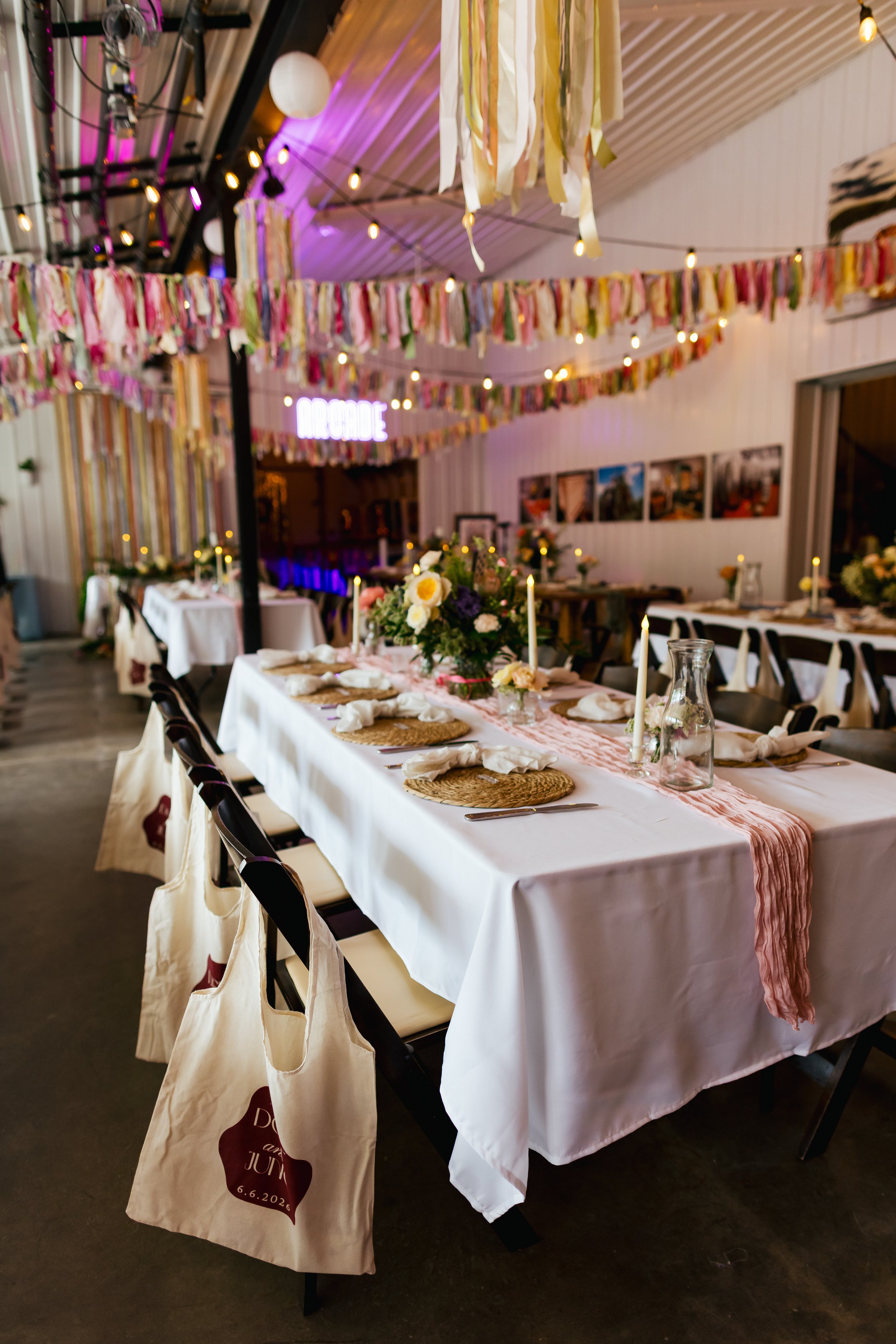 Decorated wedding reception area with a long table covered with a white tablecloth, floral centerpieces, candles, plates, and napkins. Colorful streamers and string lights hang from the ceiling, creating a festive atmosphere.