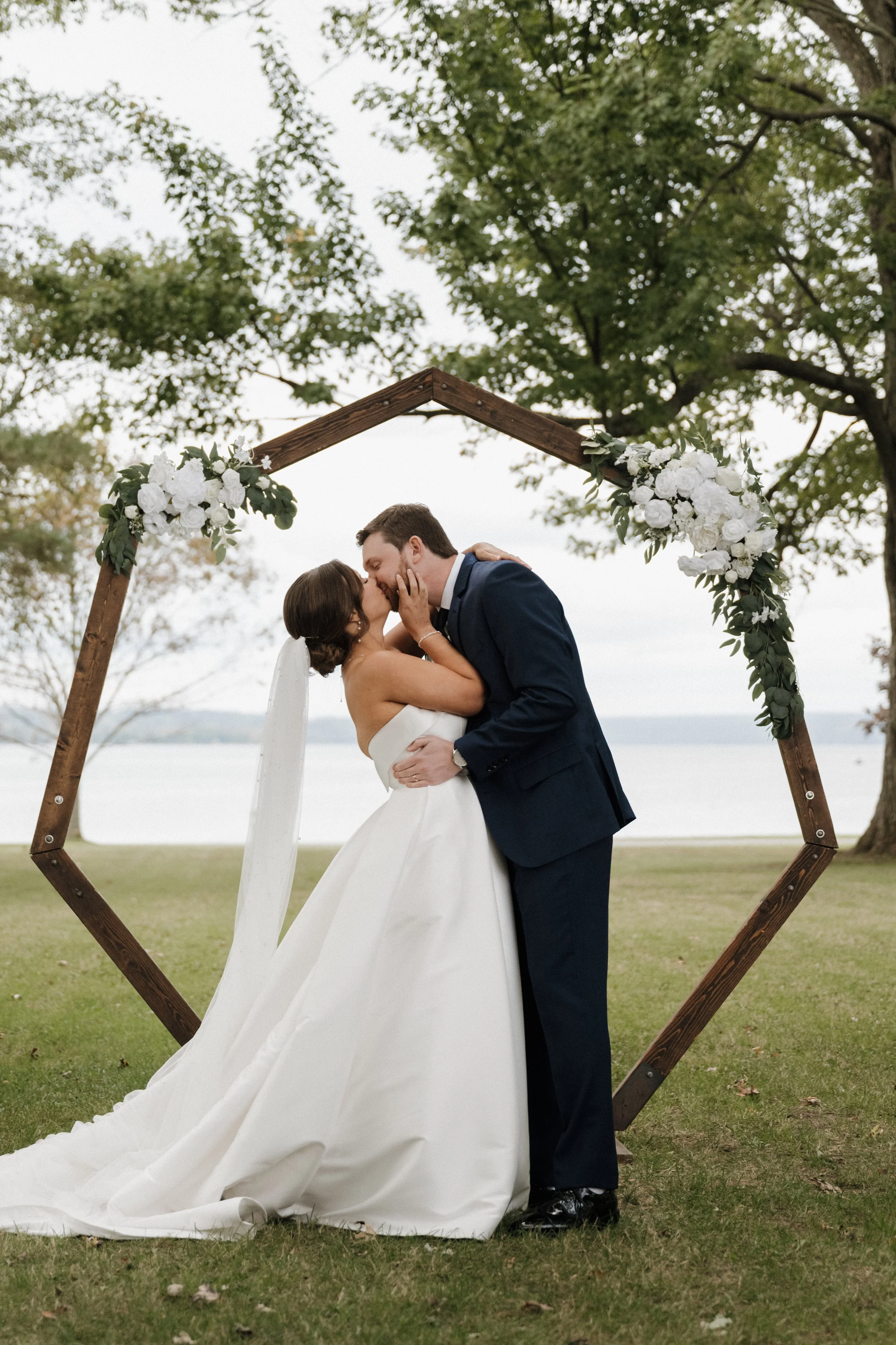 A bride and groom sharing a kiss under a wooden wedding arch decorated with white flowers and green foliage, outdoors by a lakeside with trees in the background.