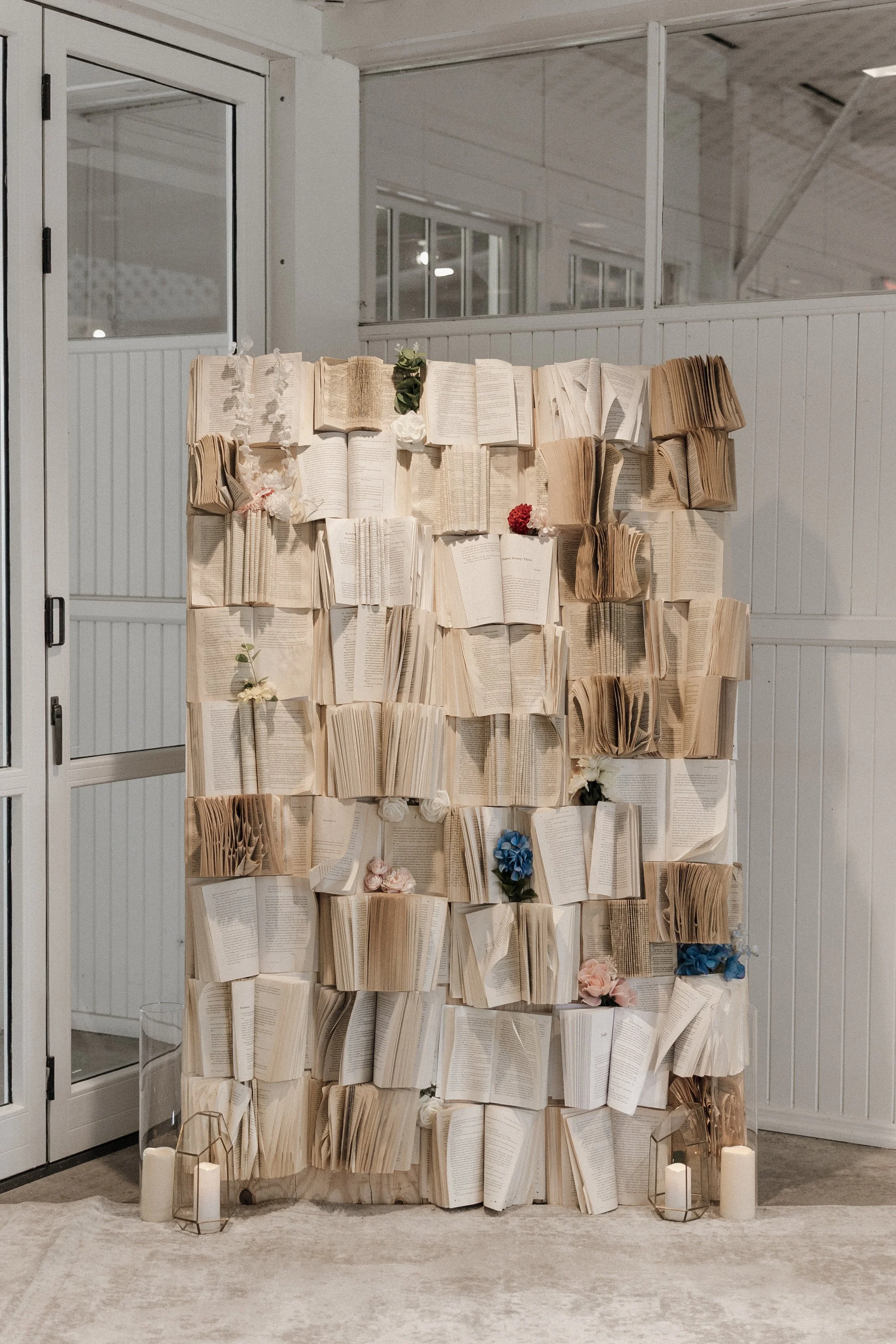 An indoor altar made from open books arranged in tiers, decorated with flowers and candles, set against white panel walls.