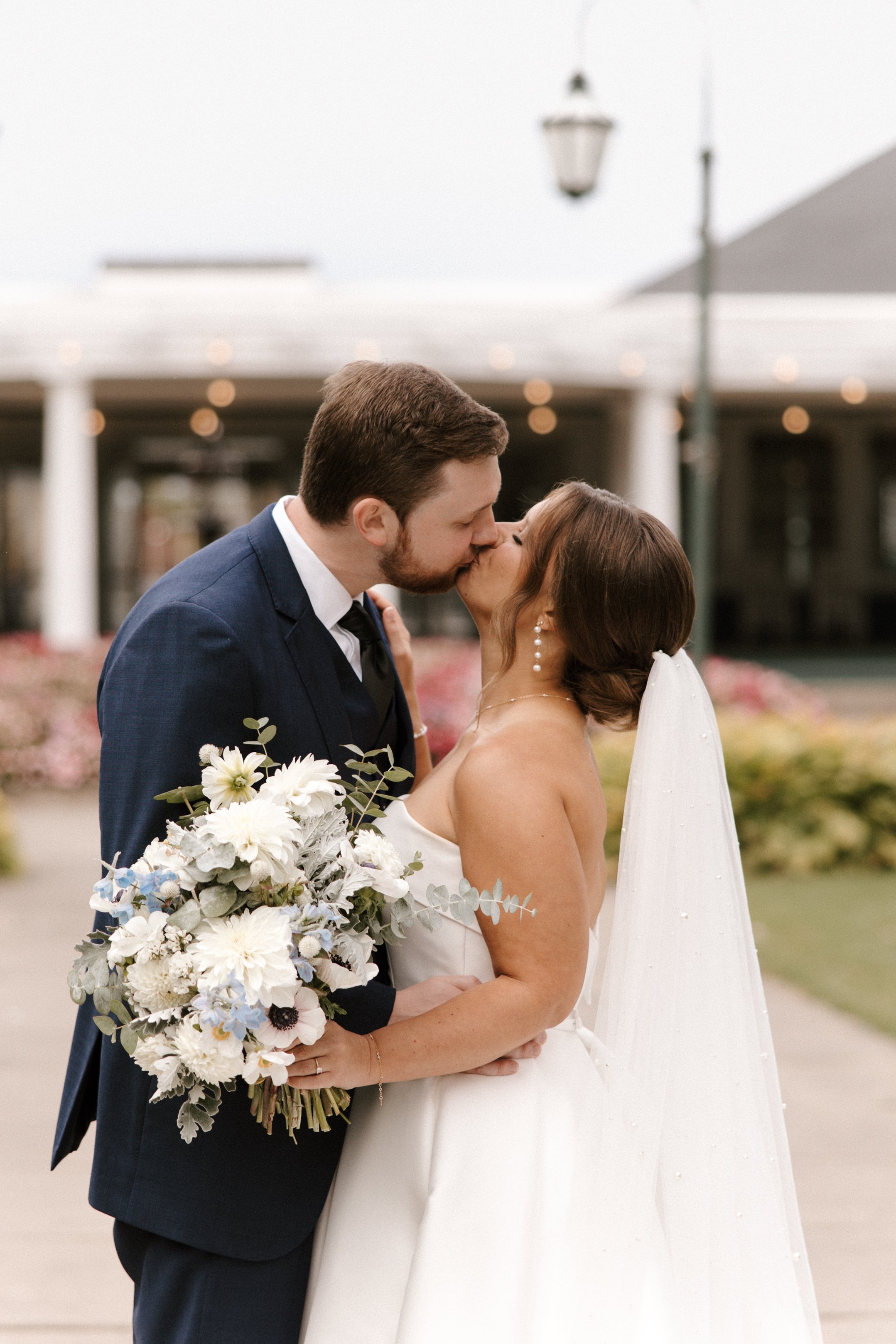 A newlywed couple shares a kiss outdoors, with the groom holding a large bouquet of white and pale blue flowers, against a background of a building and a vintage streetlamp.