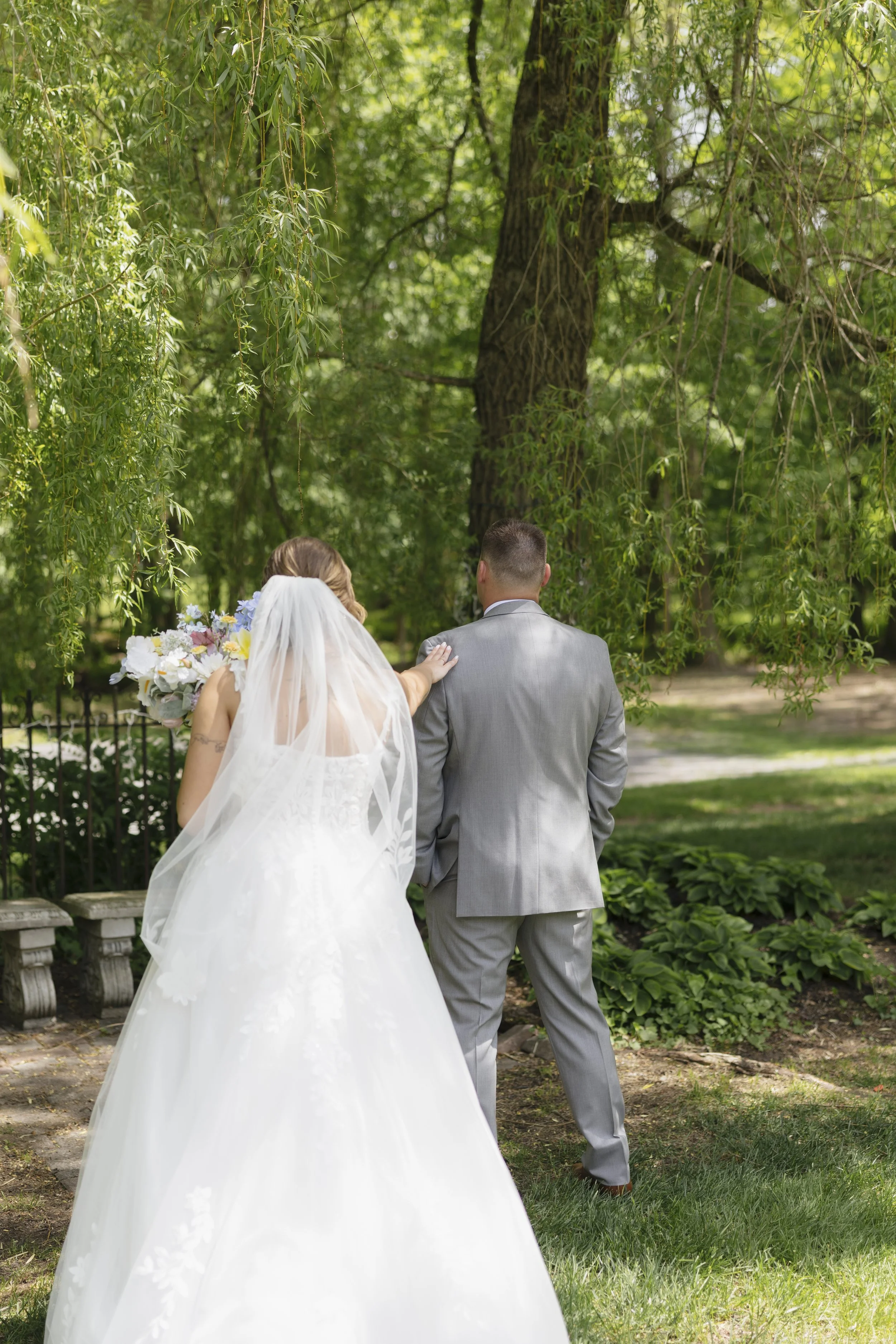 A bride and groom walking together outdoors in a park, with the bride wearing a white wedding dress and veil holding a bouquet of flowers, and the groom in a gray suit, surrounded by green trees and foliage.