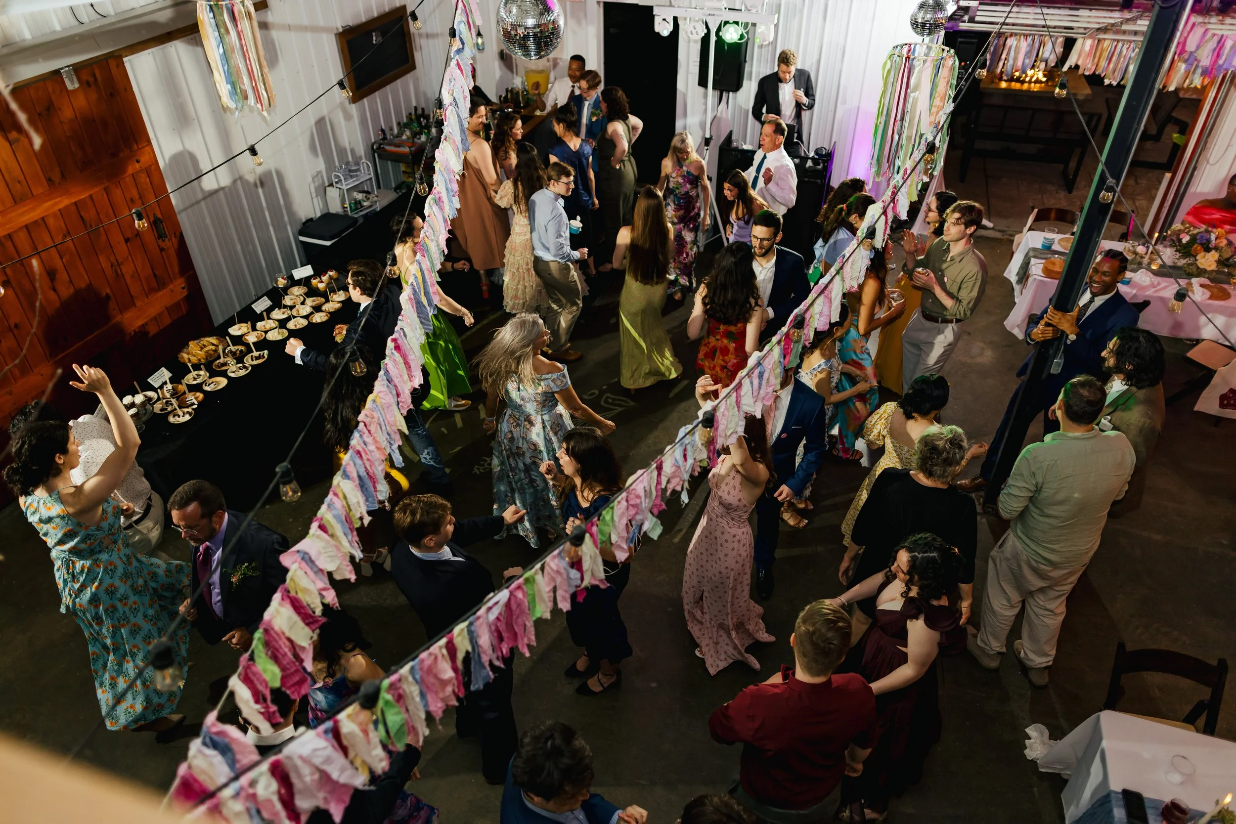 A lively indoor party or celebration with many people dancing and socializing. The room is decorated with colorful streamers and string lights, and a disco ball hangs from the ceiling. There are tables with food and drinks along the sides of the room