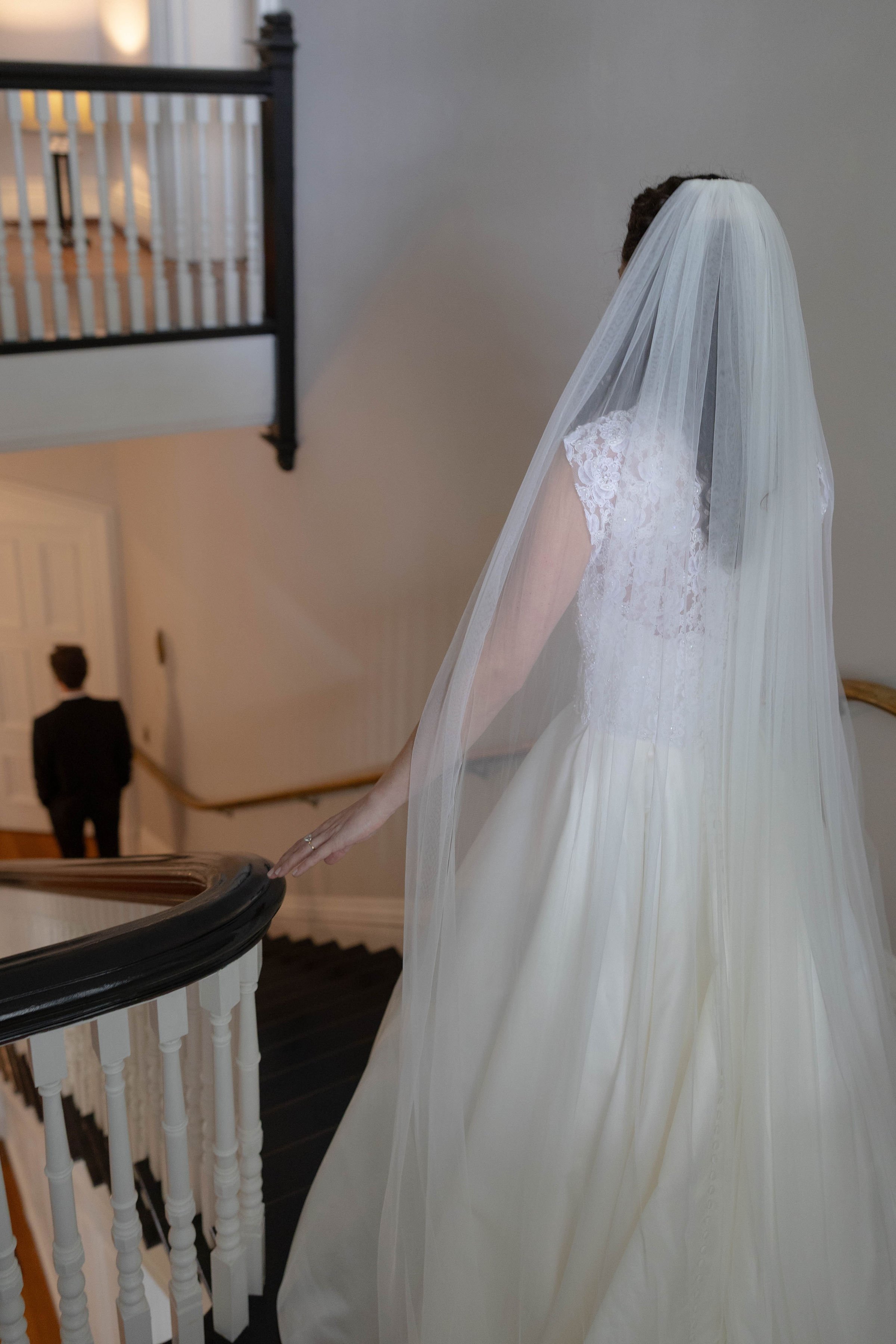 Bride in a white wedding dress with lace details and a long veil walking up a staircase, with a man in a black suit and tie in the background.