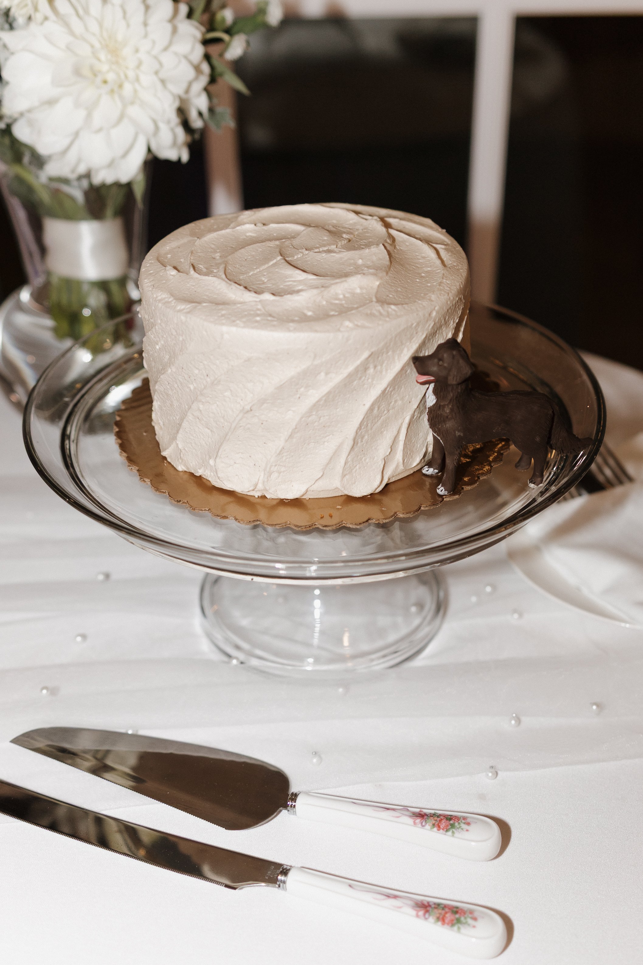 A slice of white cake with cream frosting on a glass cake stand, decorated with thinly sliced almonds on top. There is a small toy dog figurine next to the cake. In the background, there is a vase with white flowers on a table.