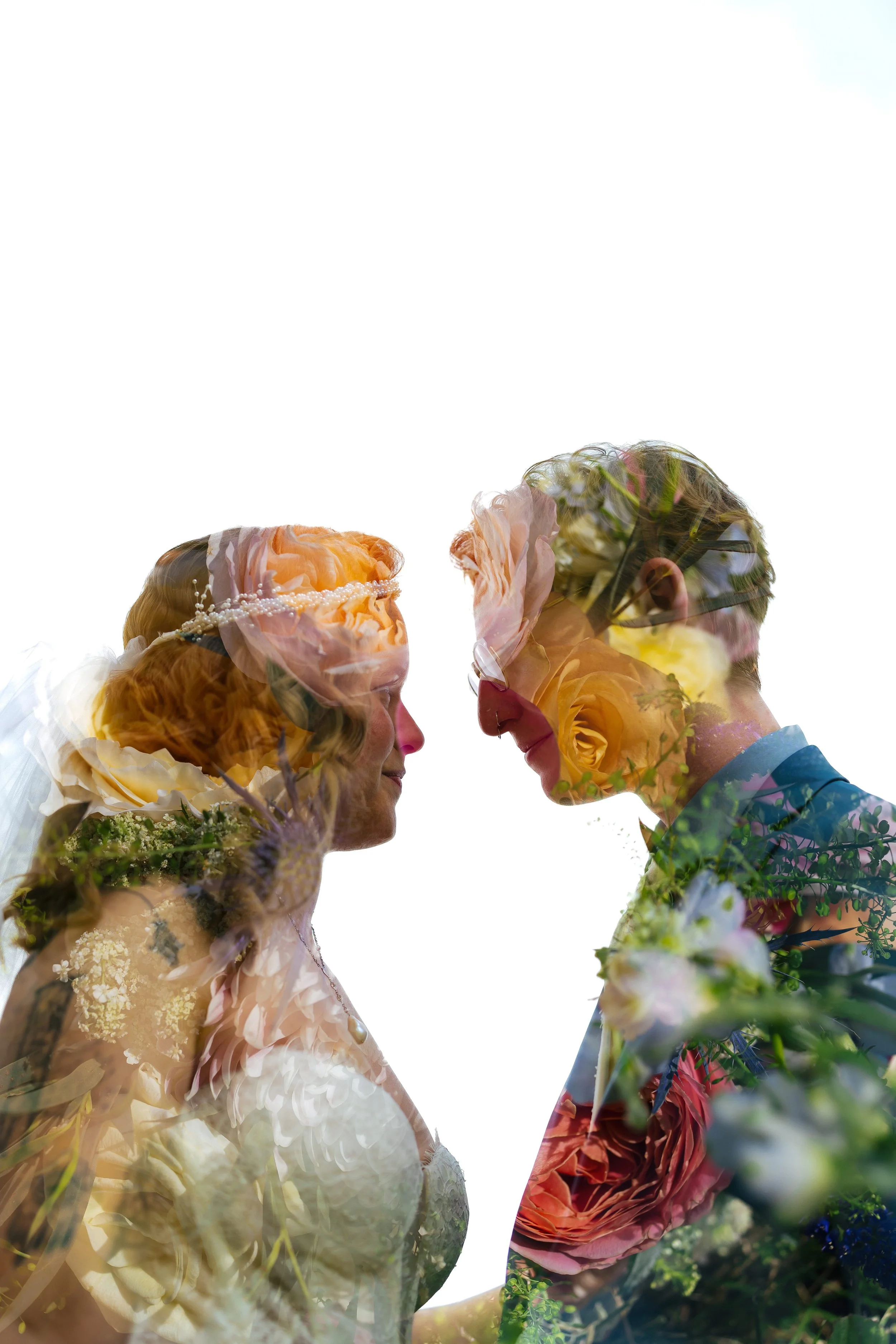 A couple, a woman with red hair and a man with glasses, facing each other closely, surrounded by flowers, with their faces close together, overexposed sky in the background.