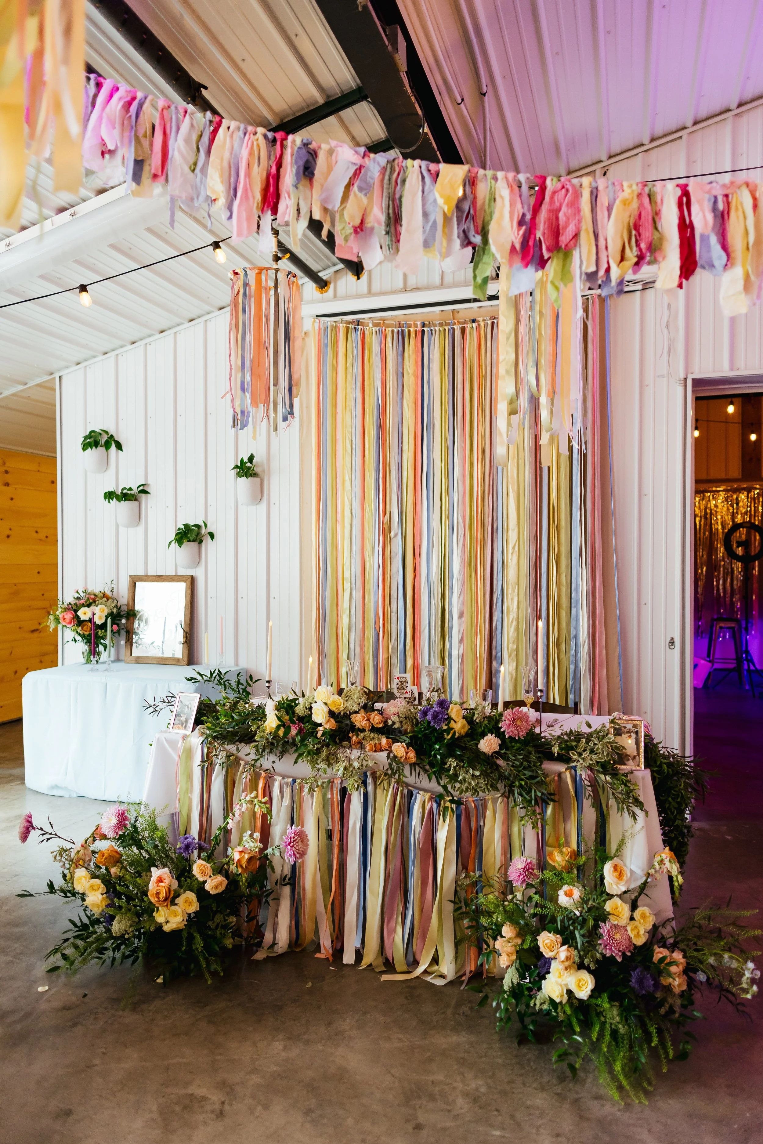 Colorful ribbon decorations and floral arrangements decorate a table for a celebration inside a venue with white and wood-paneled walls.
