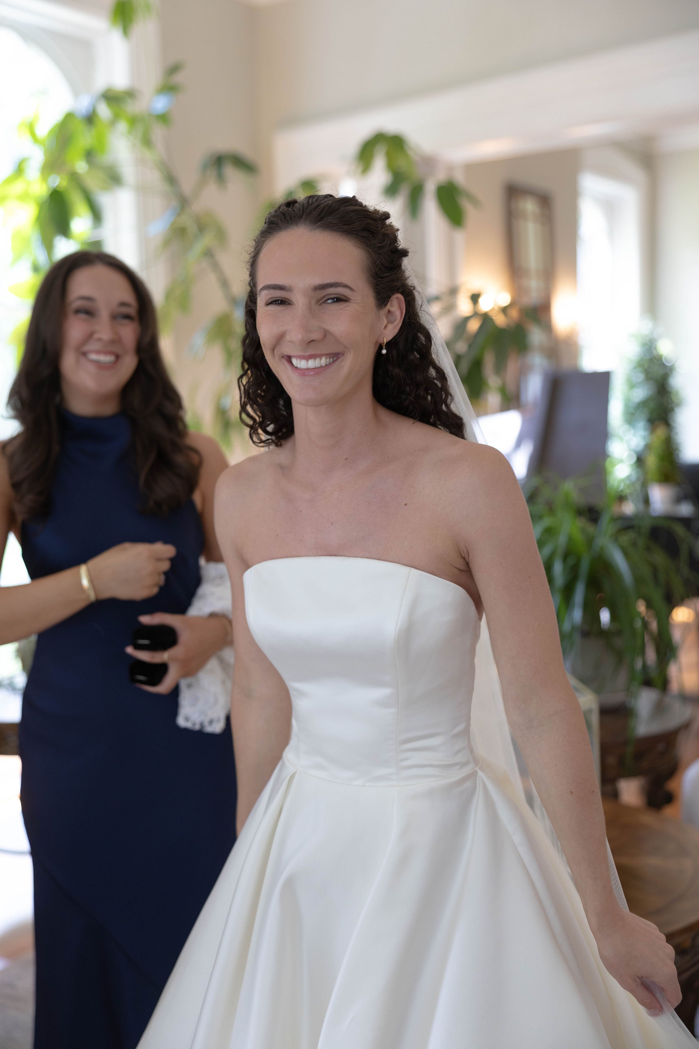 A bride smiling in a strapless white wedding gown, with a woman in a navy dress standing behind her in a brightly lit room with green plants.