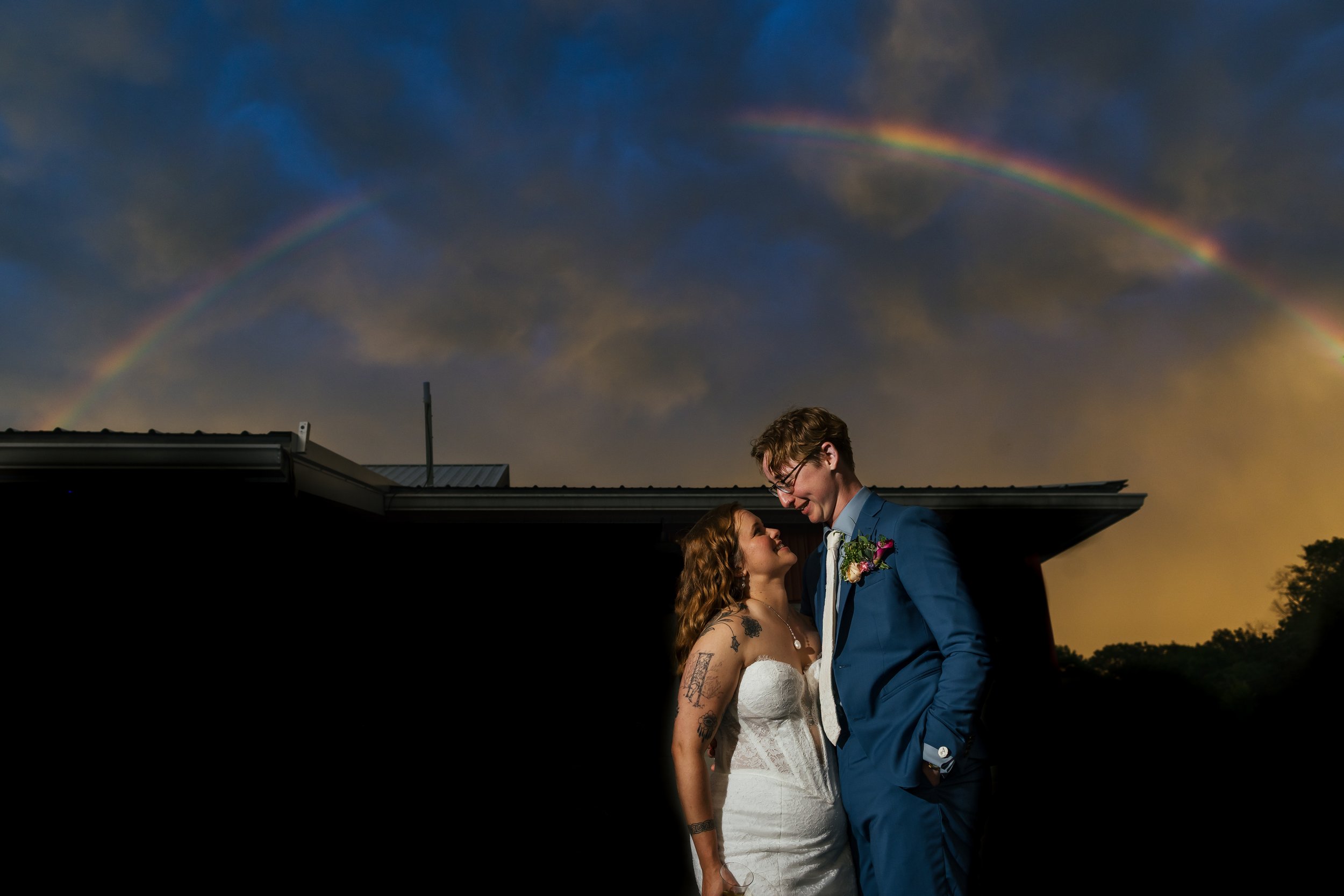 A couple, dressed in wedding attire, look at each other affectionately outdoors at sunset under dark clouds with a rainbow in the sky.