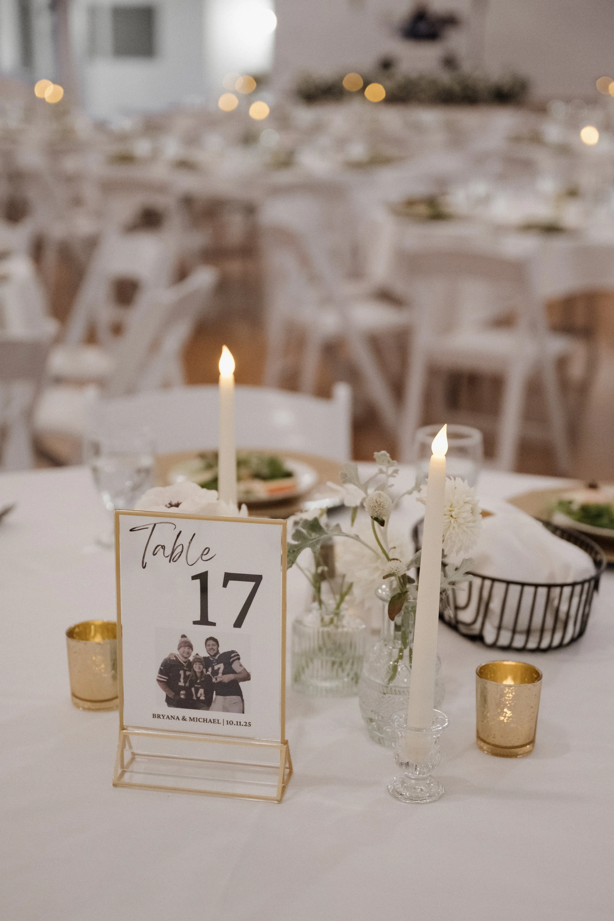 A table arrangement at a wedding or celebration with a framed photo of a sports team, candles, flowers, and gold votive holders. The background shows more tables with similar settings and a blurred area.